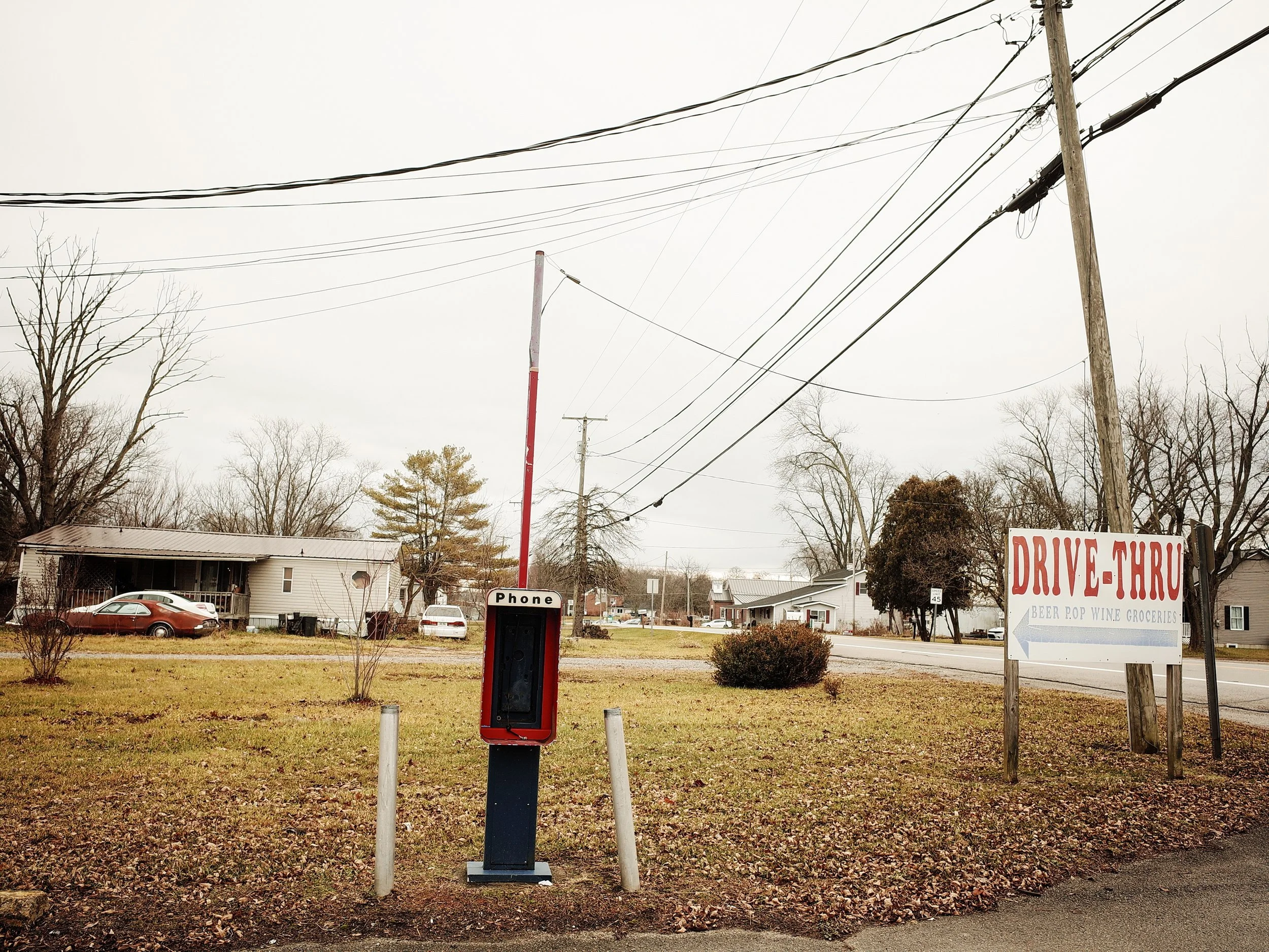 A color documentary photograph of an isolated red public payphone booth standing in a grassy yard next to a drive-thru sign in Lynchburg, Ohio, from One Way Out, Lord by Parker J Pfister.