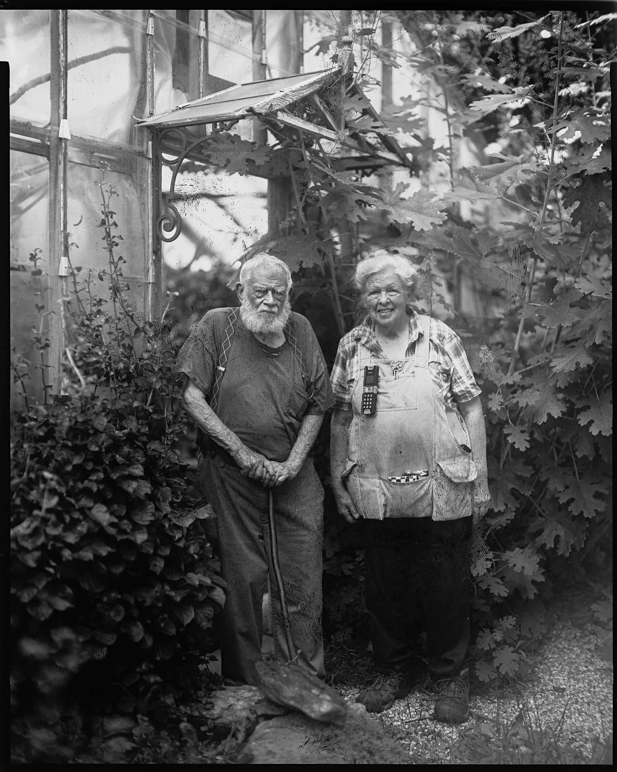 A full-length black and white portrait of an elderly man with a shovel and a woman in overalls standing outside a rustic glass greenhouse surrounded by lush foliage in Appalachia. Large format photography