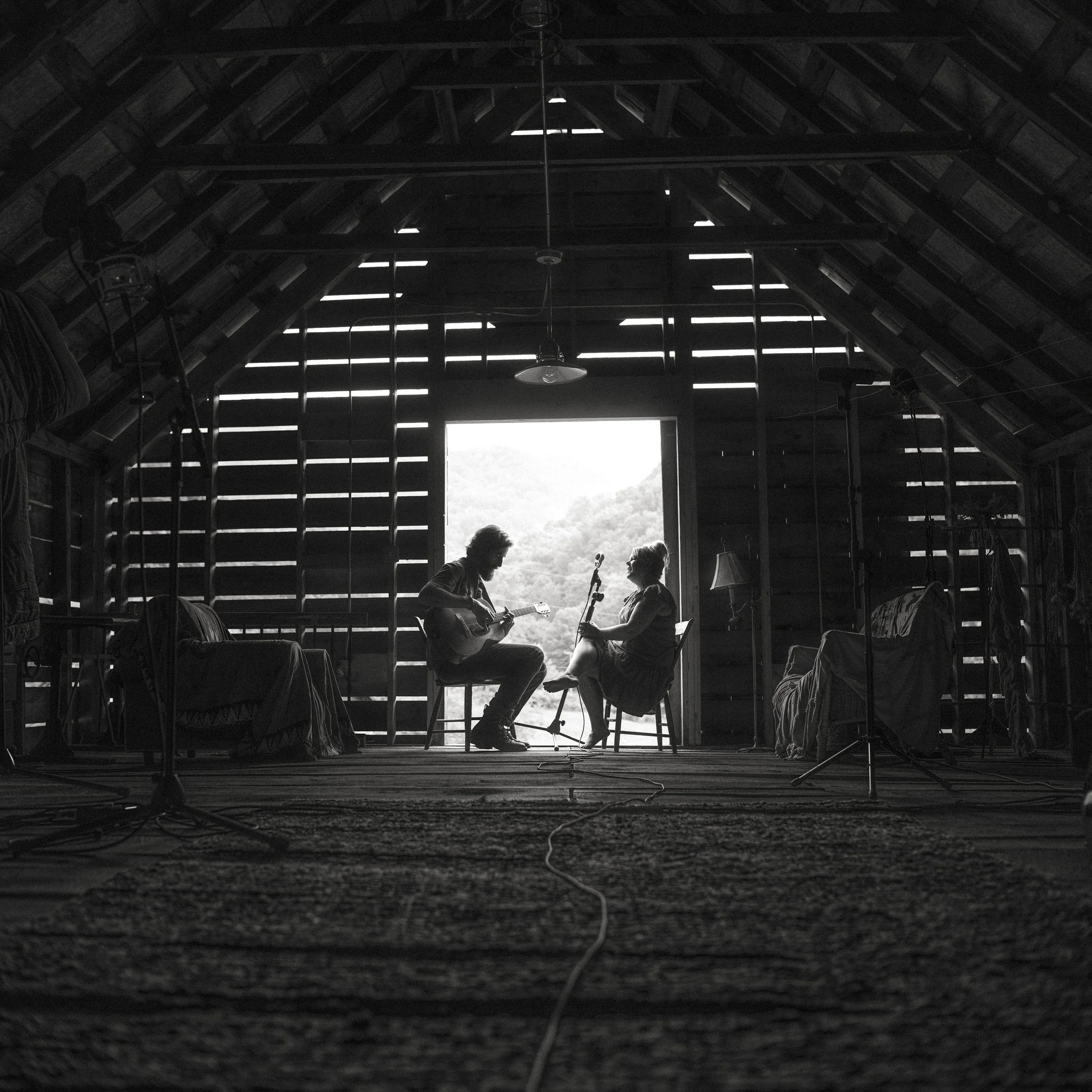 Silhouetted interior view of musicians Tyler Ramsey and Donna Ray Norton playing acoustic guitars inside a rustic barn opening at Rare Bird Farm in Madison County, NC while recording their album of Appalachian ballads, photographed by Parker J. Pfist