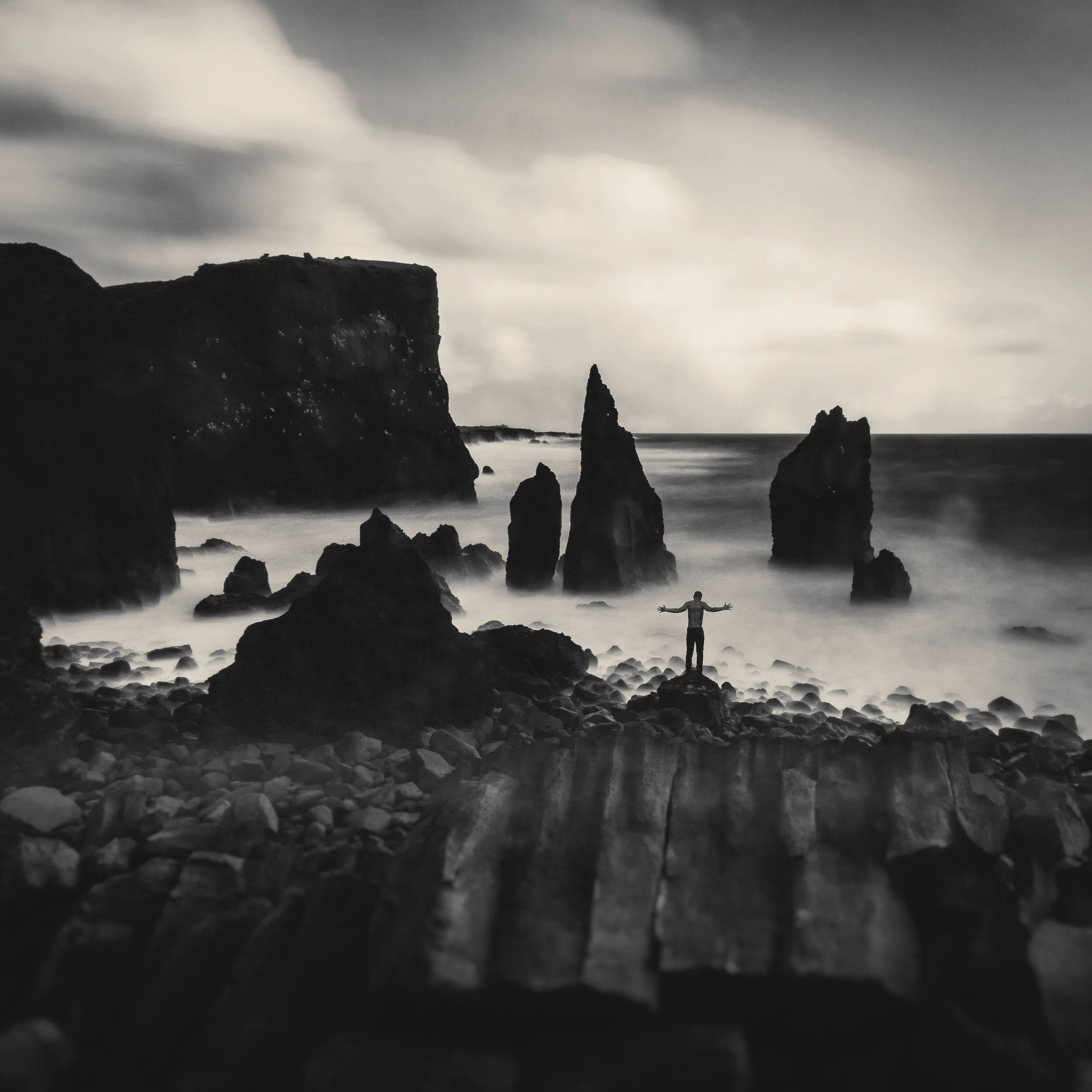 A dramatic black and white fine art photograph capturing a lone figure standing with outstretched arms on a dark, rocky shore in Iceland, surrounded by towering sea stacks and crashing waves under an overcast sky.