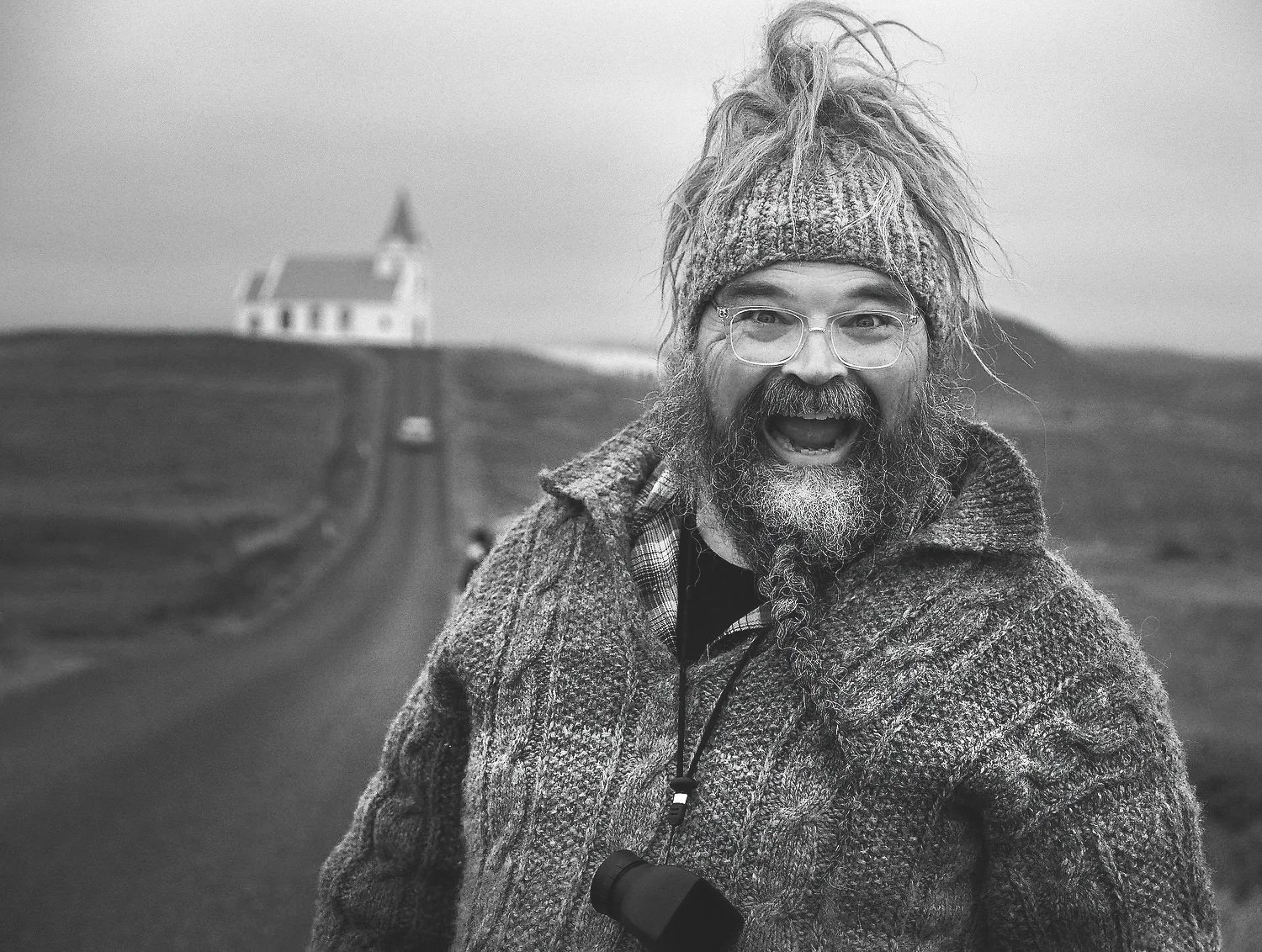 Expressive, joyful black-and-white portrait of photographer Parker Pfister wearing round glasses and a textured knit beanie with long dreadlocks, smiling widely in front of a winding road leading to a classic white Icelandic church with a steeple