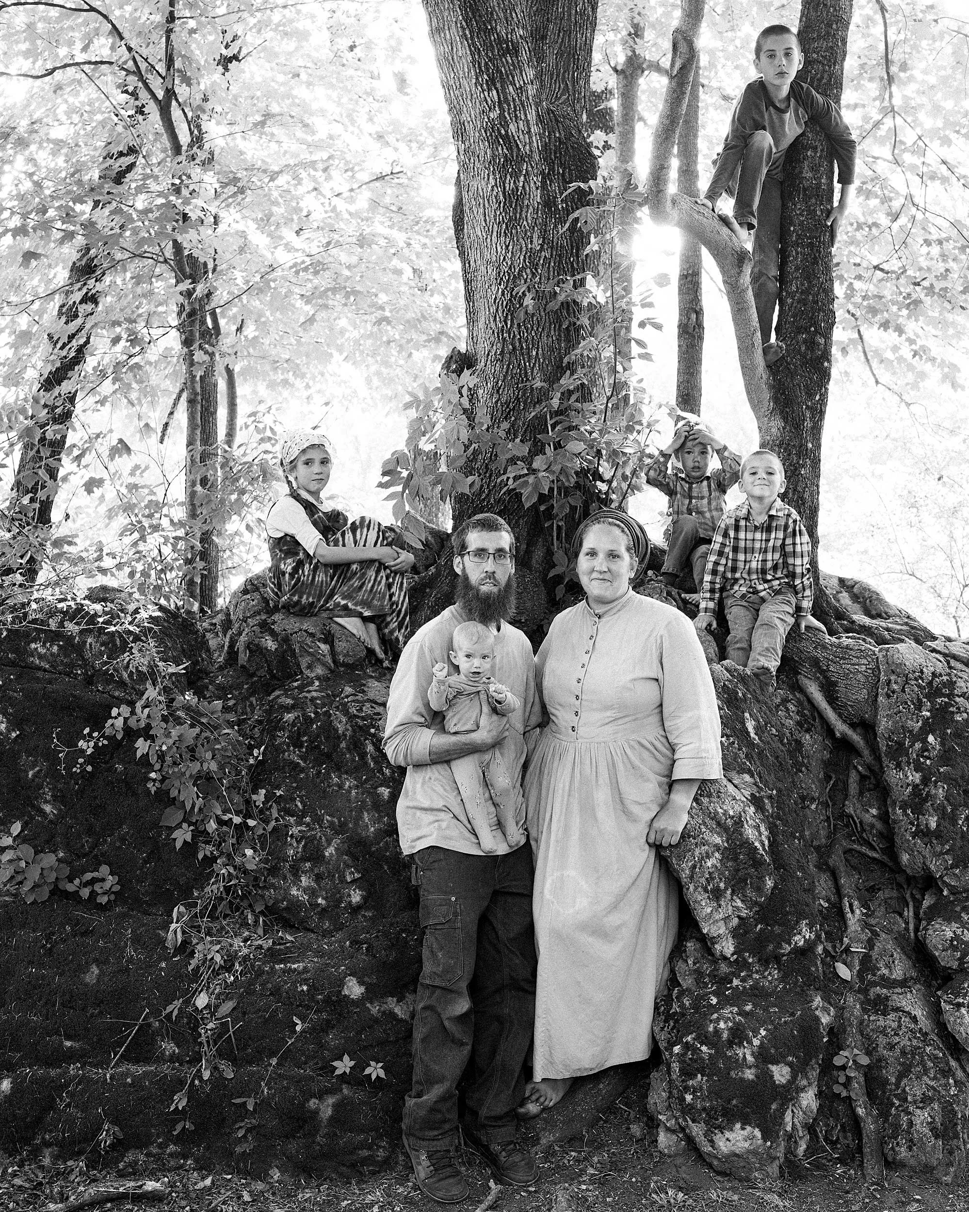 Black and white documentary portrait of a mountain family—a mother and father holding a baby, with three older children perched above them on massive, exposed tree roots in a Western North Carolina forest.