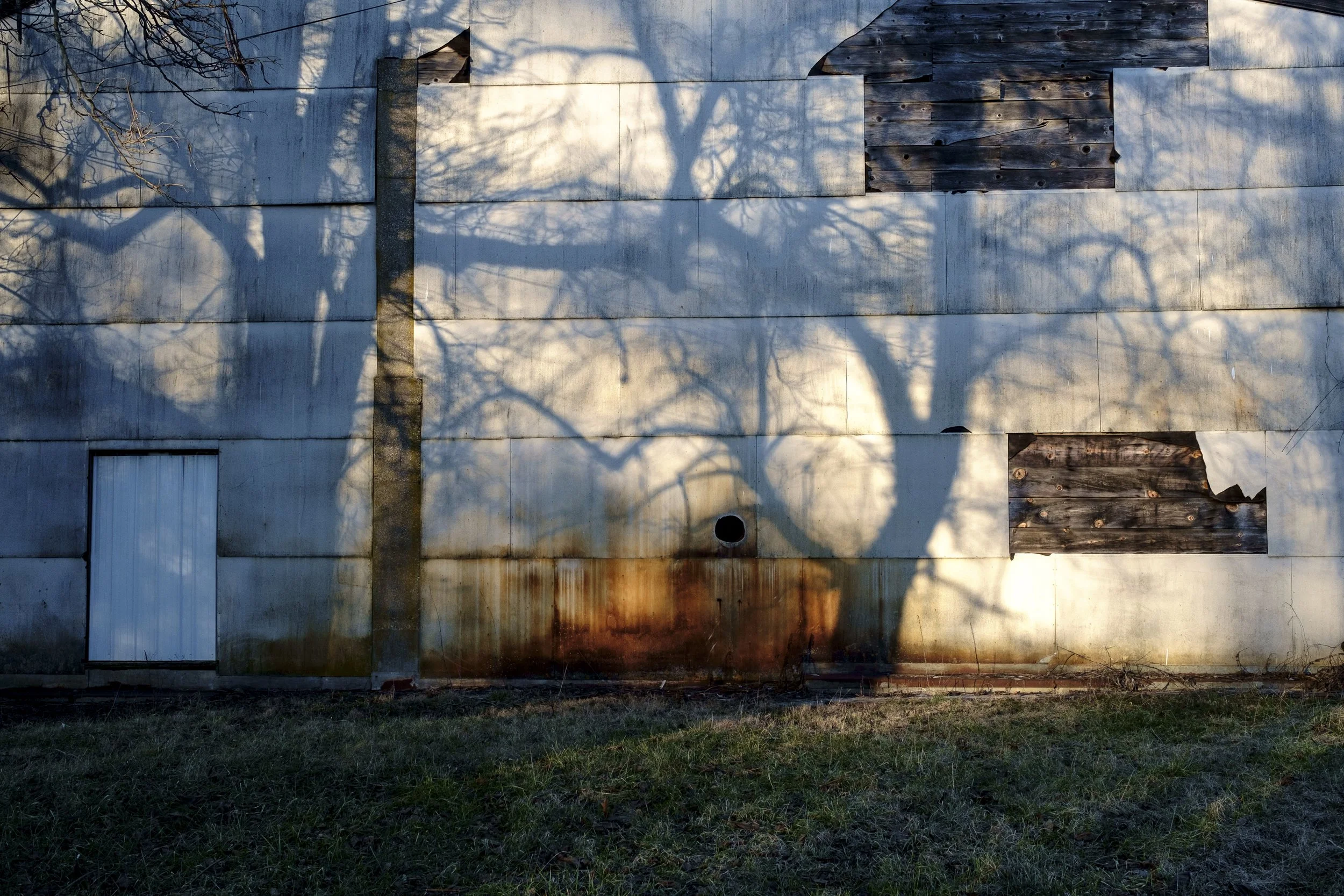 A color documentary photograph of dark, bare tree branch shadows cast across the weathered white siding and boarded-up windows of an old building, from One Way Out, Lord by Parker J Pfister.