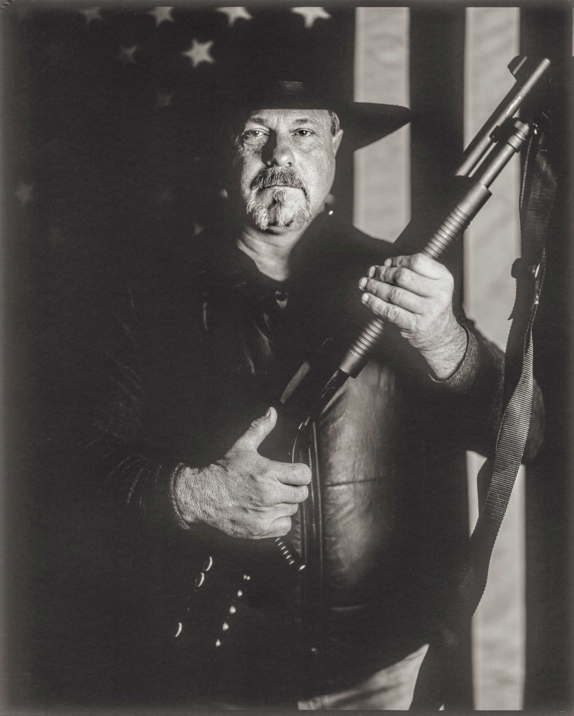 A powerful black and white portrait of a man wearing a cowboy hat and leather vest, holding a shotgun across his chest against an American flag backdrop, capturing a face of the mountain community in Appalachia.