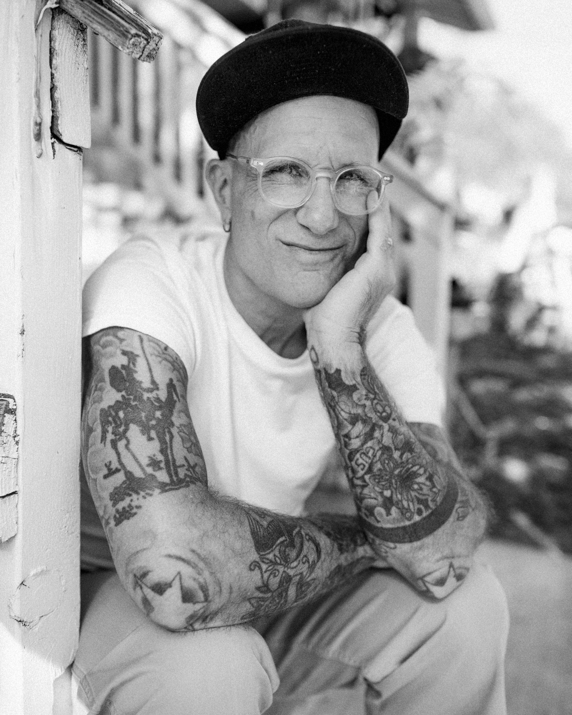 Black and white portrait of musician Gary Jules resting his chin on his hand in a cap, glasses, and white t-shirt, photographed by Parker J. Pfister.