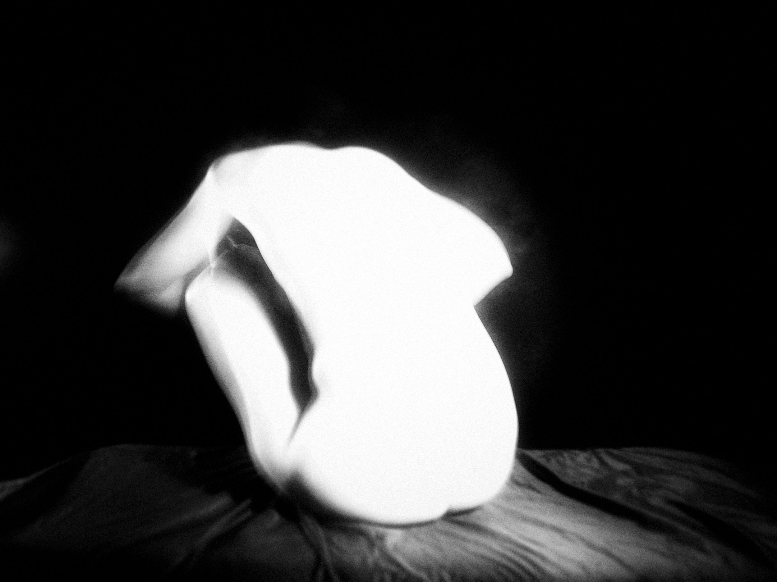 A high-contrast monochrome fine art nude figure study captured with an intentional vintage lens blur and oil filter, creating a brilliant white glow around the form of a seated figure.