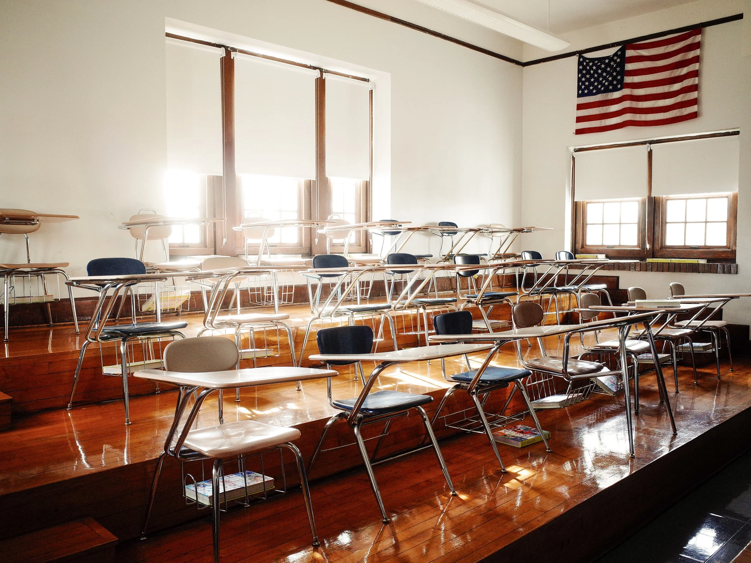 A color documentary photograph of an empty physics classroom with tiered wooden floors, rows of school desks, and an American flag under bright window light in Greenfield, Ohio, from One Way Out, Lord by Parker J Pfister.