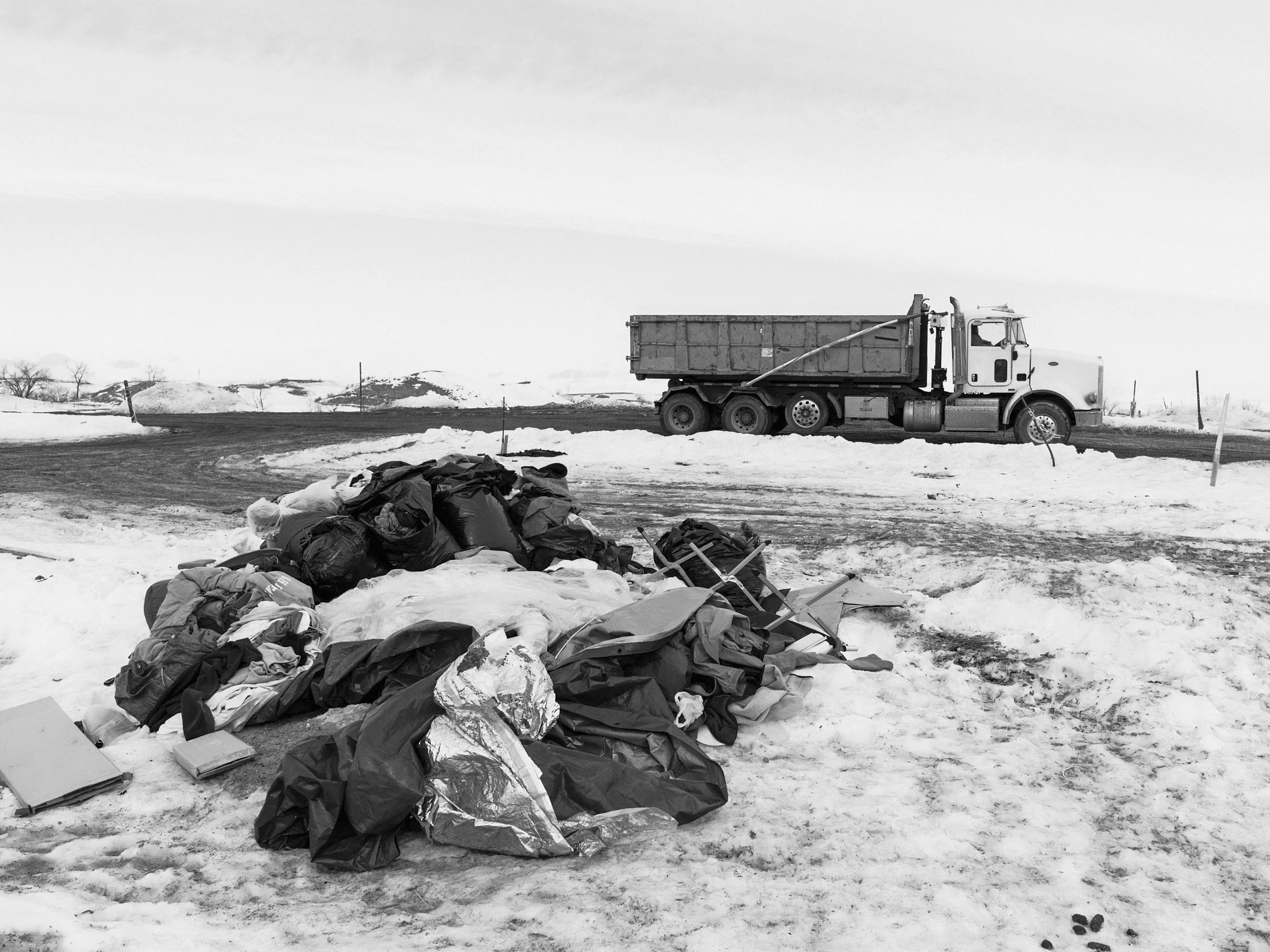 Black and white documentary photograph of a large pile of discarded tarps, gear, and debris sitting on a patch of snowy, muddy ground at the Standing Rock camp, with a large dump truck parked in the background.