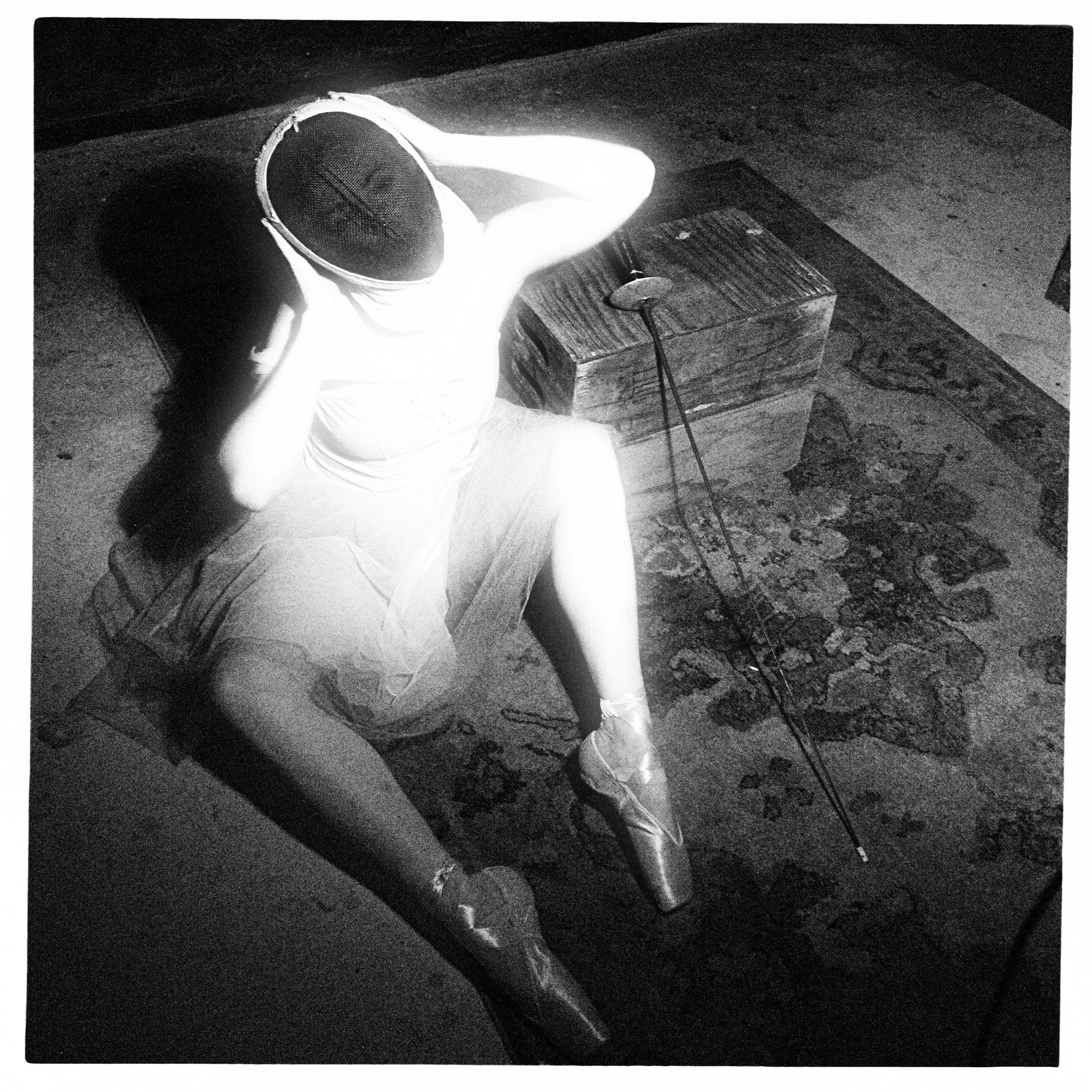 A high-contrast fine art photograph taken from an overhead perspective, showing a ballerina in a tutu and pointe shoes seated on a patterned rug, adjusting a dark fencing mask next to her foil.