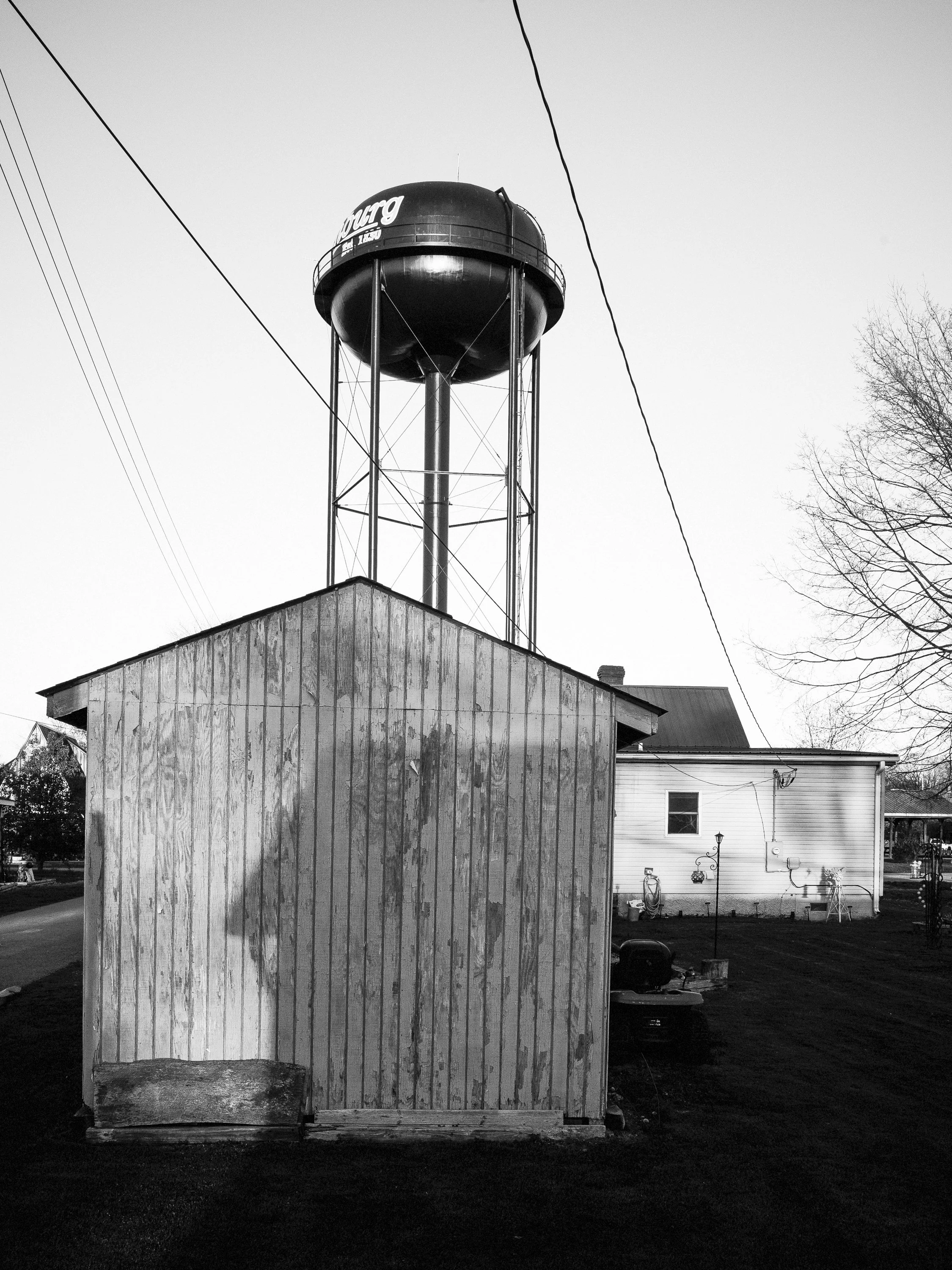 A vertical black and white documentary photograph of a weathered wooden shed with a large municipal water tower rising directly behind it into the sky, from One Way Out, Lord by Parker J Pfister.