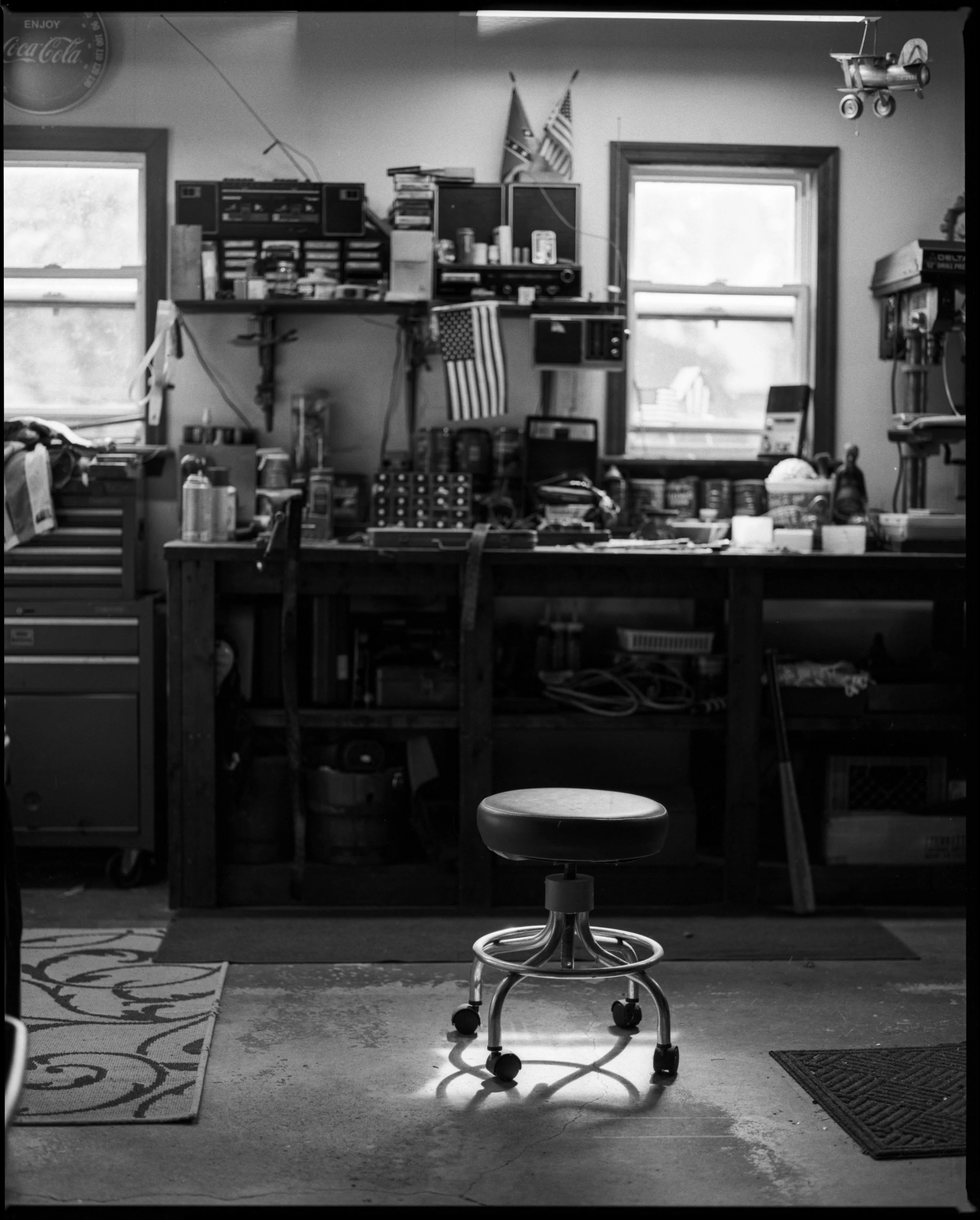 A high-contrast black and white photograph of an empty garage workshop, featuring a rolling stool under a sharp pool of light in front of a detailed, tool-filled workbench, from One Way Out, Lord by Parker J Pfister.