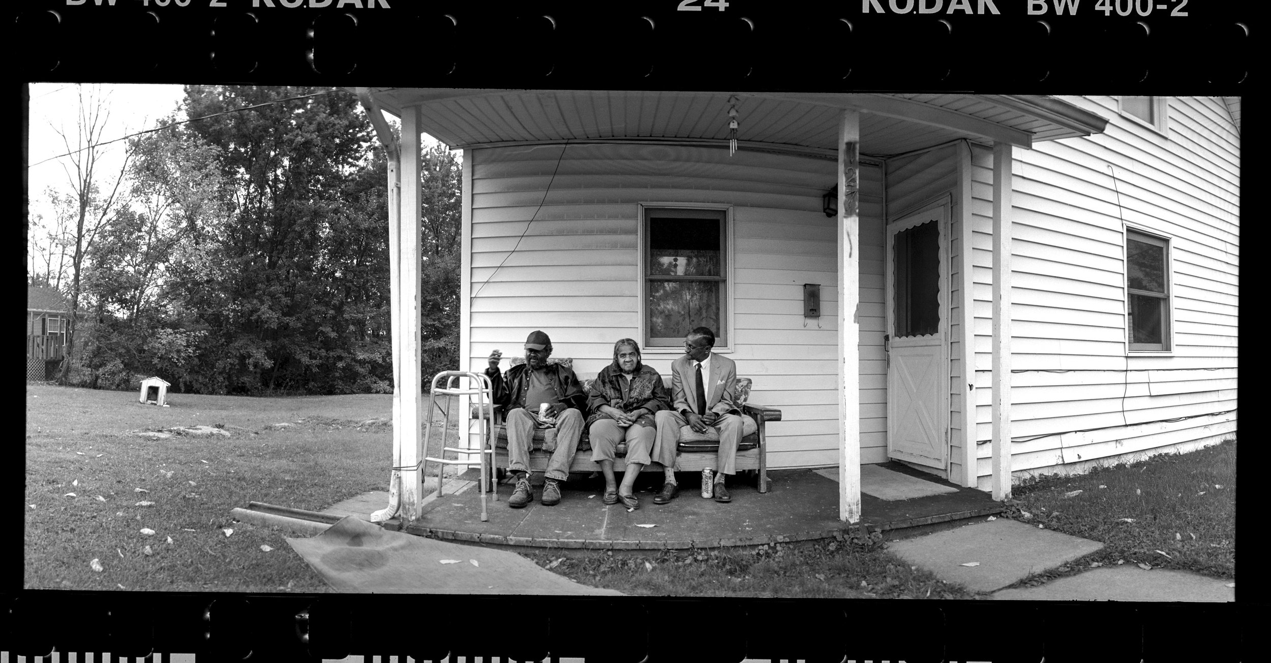 A panoramic film black and white documentary photograph of three people sitting and talking on a wooden bench on the porch of a white residential house, from One Way Out, Lord by Parker J Pfister.