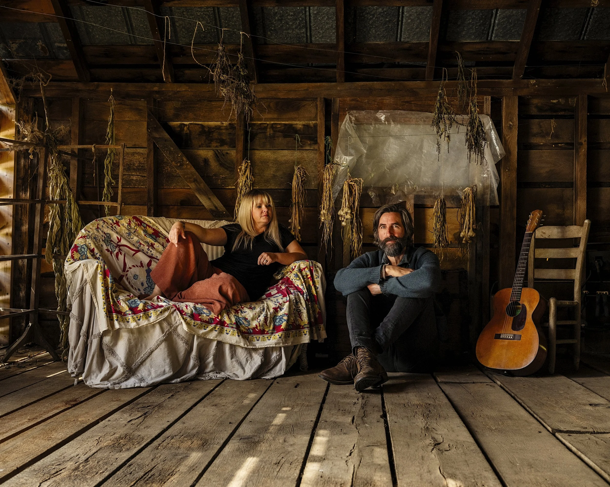 Portrait of musicians Tyler Ramsey and Donna Ray Norton seated together inside a rustic wooden barn at Rare Bird Farm in Madison County, NC with an acoustic guitar, photographed by Parker J. Pfister.