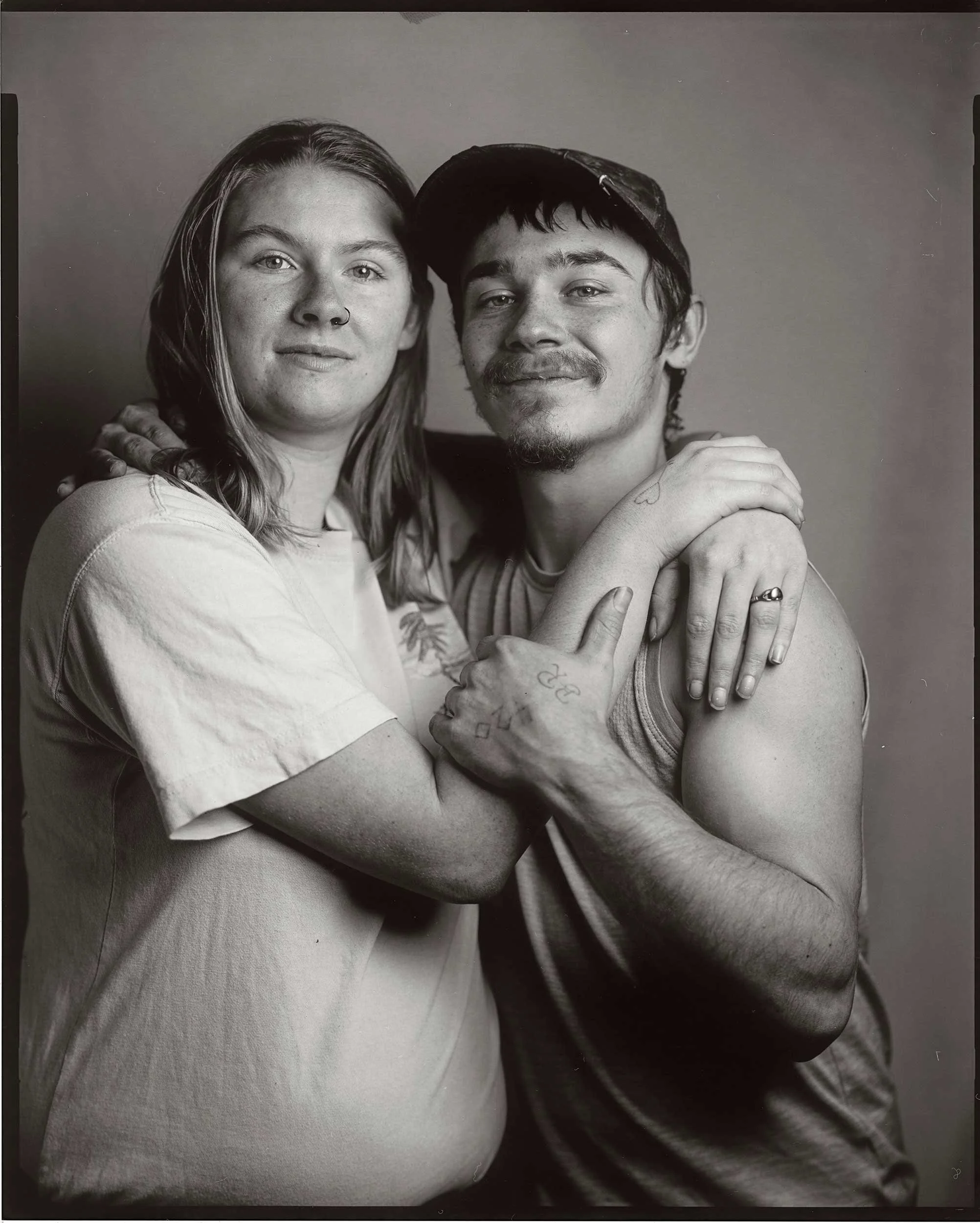 An 8x10 large format black and white paper negative portrait of a young couple smiling and embracing tightly against a plain studio backdrop at the old Depot in Marshall, North Carolina.
