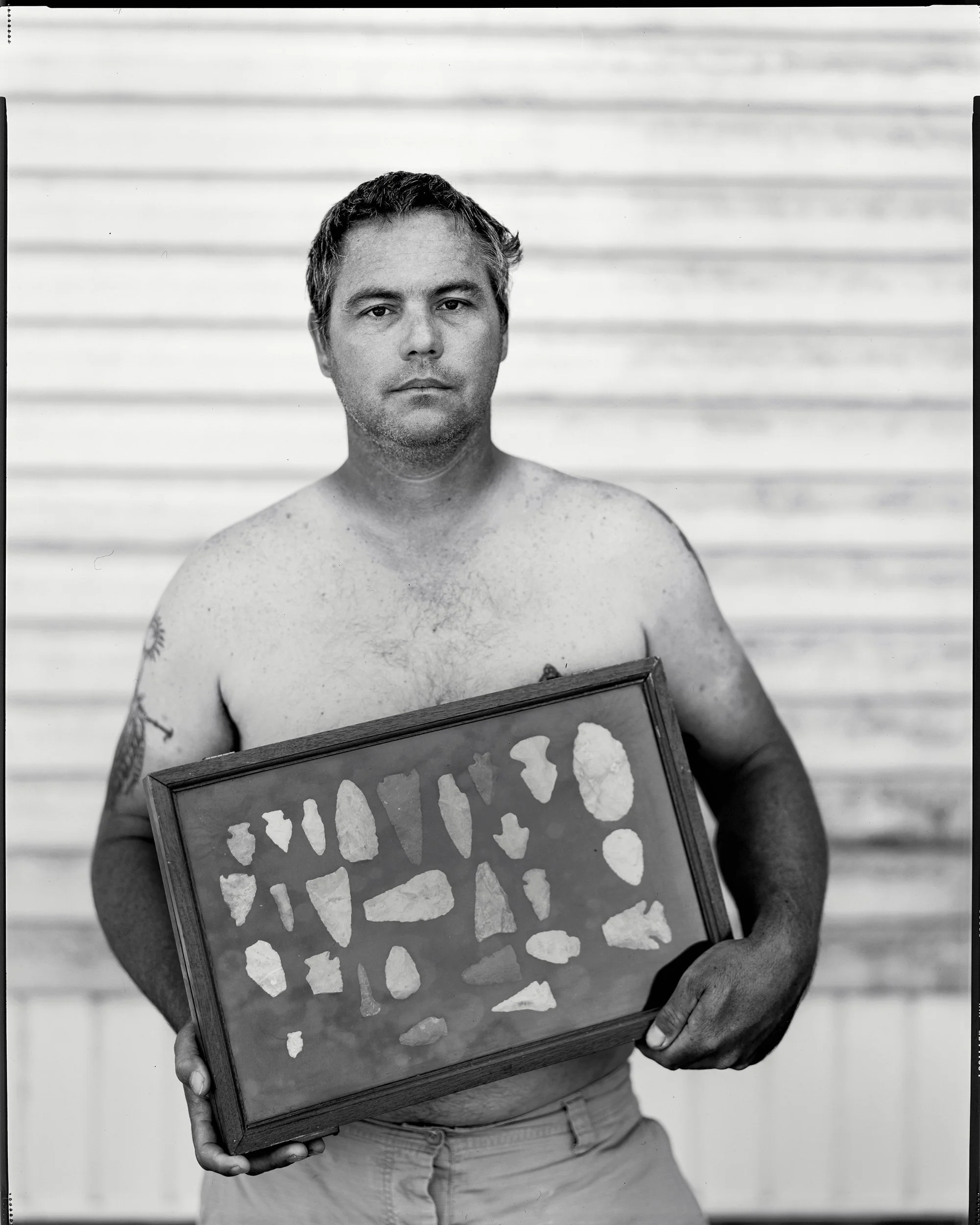 A striking black and white portrait of a shirtless man holding a framed display case of found arrowheads against a light siding background, from One Way Out, Lord by Parker J Pfister.