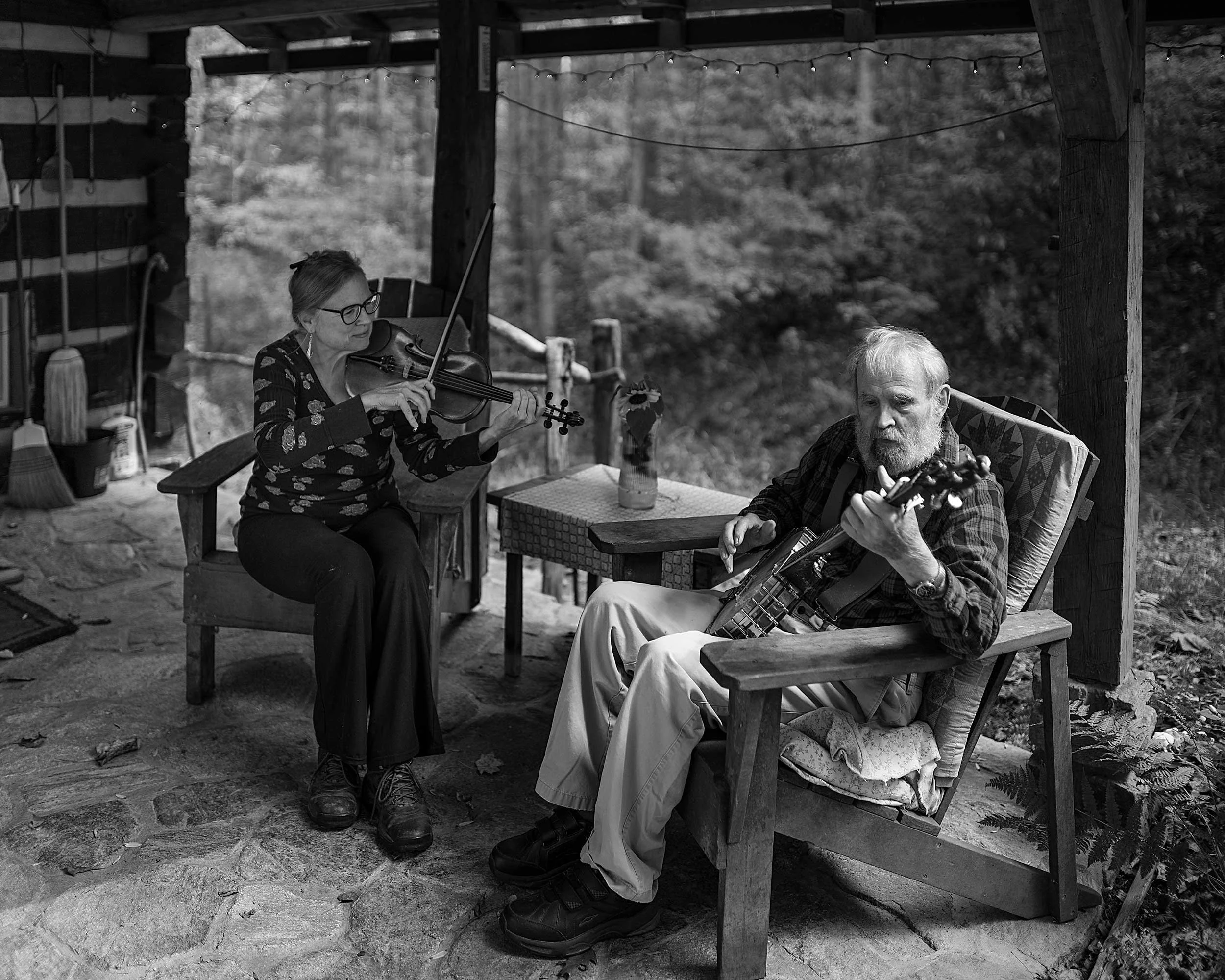 Black and white documentary photograph of a woman playing the fiddle and a man playing an acoustic instrument while sitting together on a rustic log cabin porch in Western North Carolina.