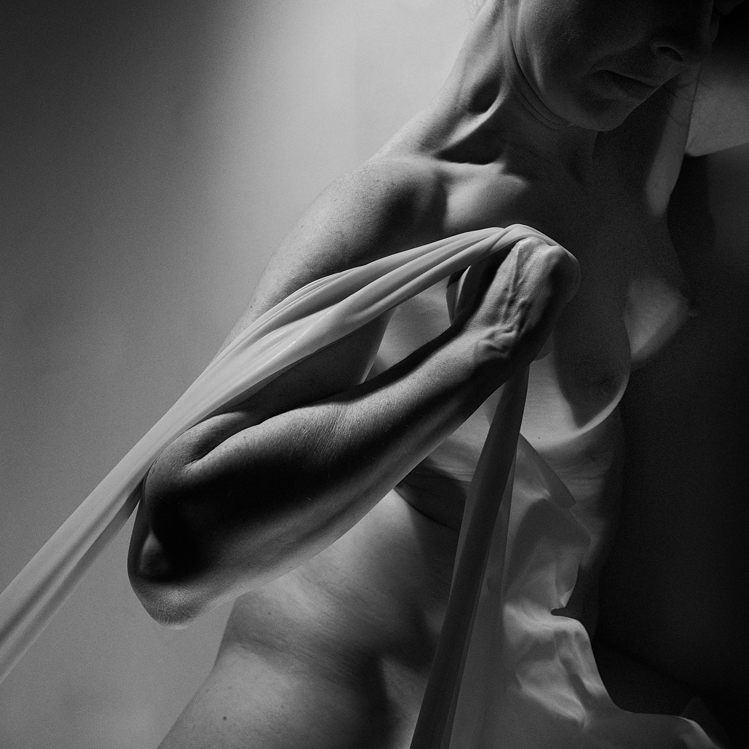 A high-contrast black and white fine art nude photograph focusing on the classic musculature and lines of a torso, draped elegantly with a long white fabric.