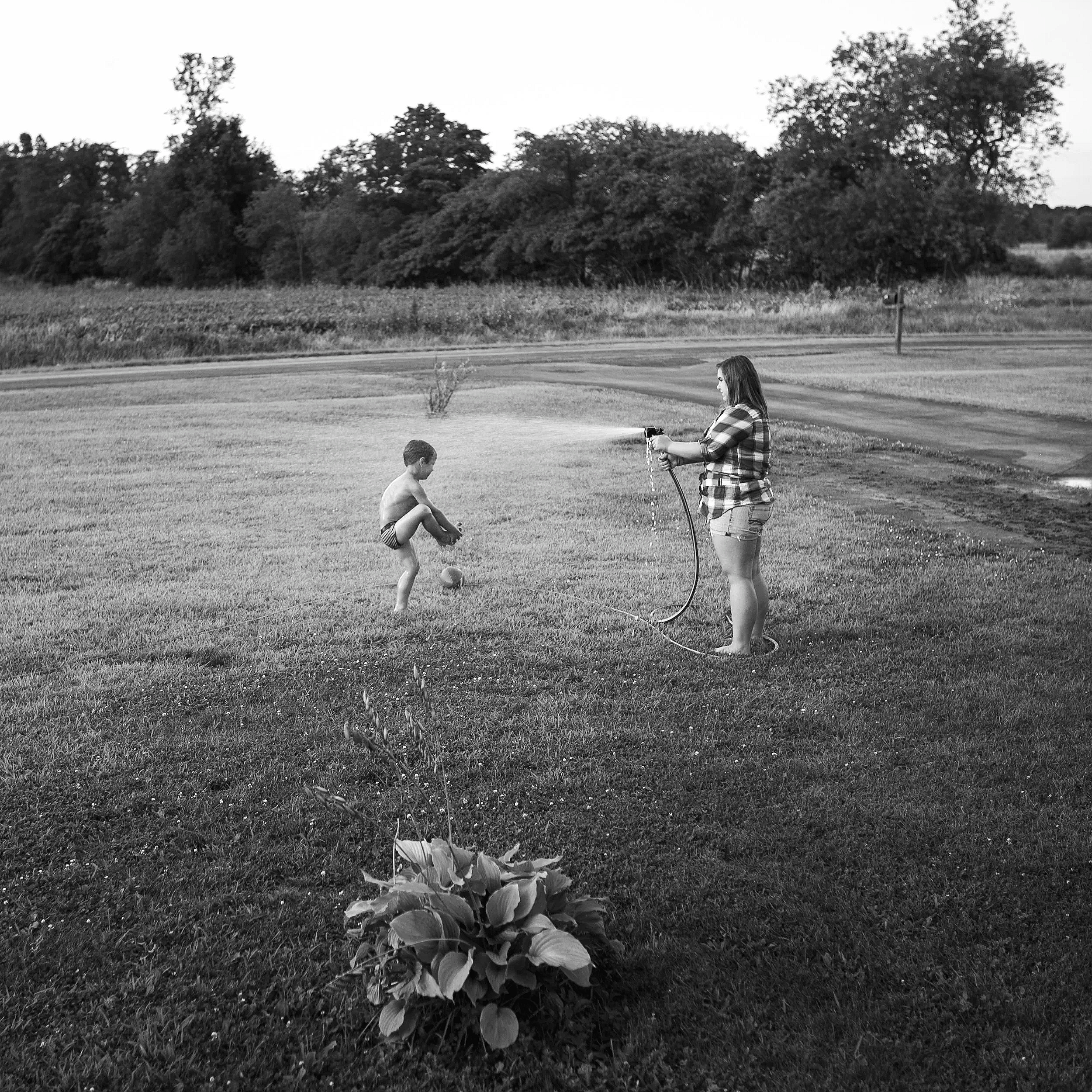 A black and white documentary photograph capturing a woman spraying a young boy with a garden hose in a wide grassy yard, from the One Way Out, Lord series by Parker J Pfister.