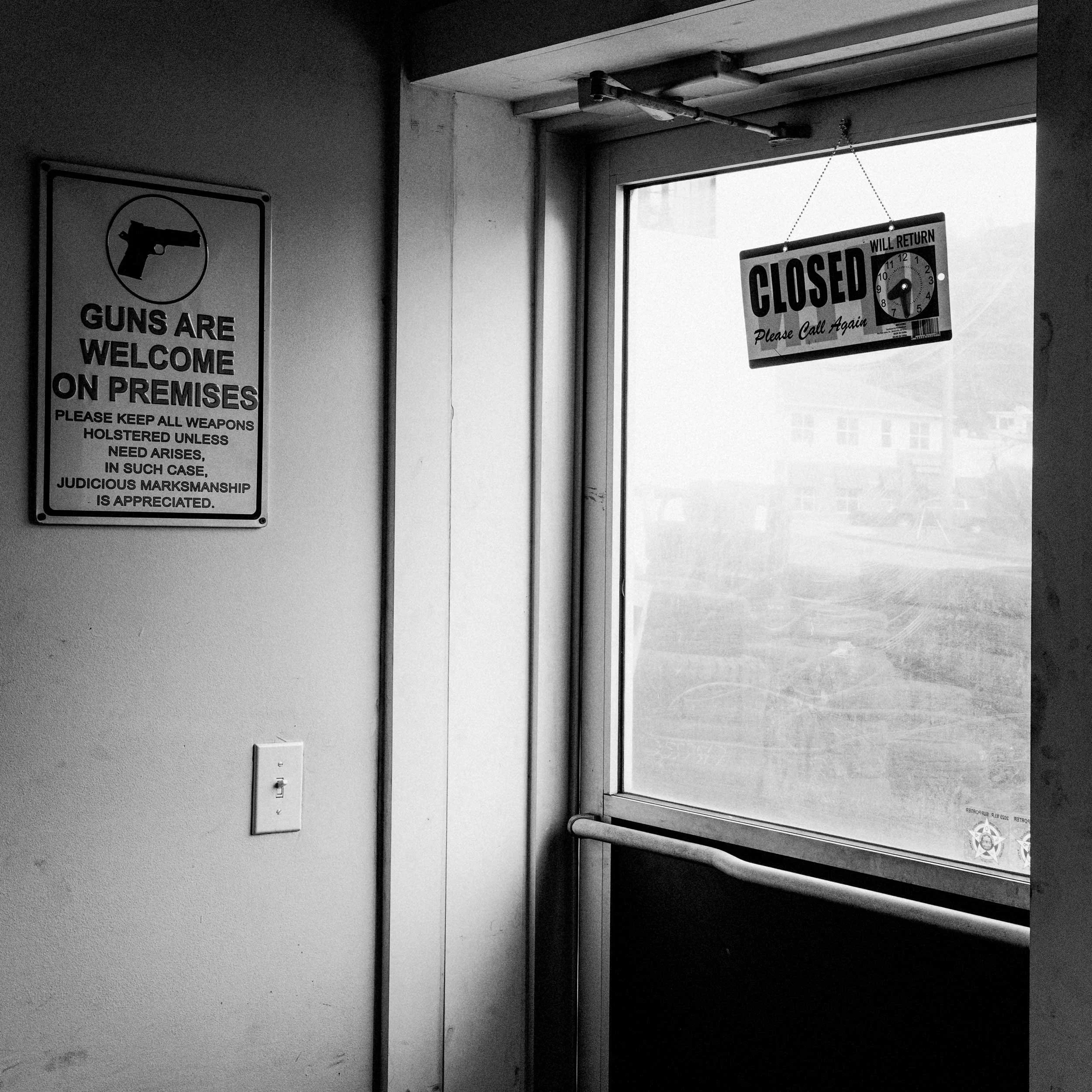 High-contrast black and white documentary photograph of a glass door with a hanging "Closed Please Call Again" sign, next to a wall sign reading "Guns Are Welcome On Premises" inside a Western North Carolina building.