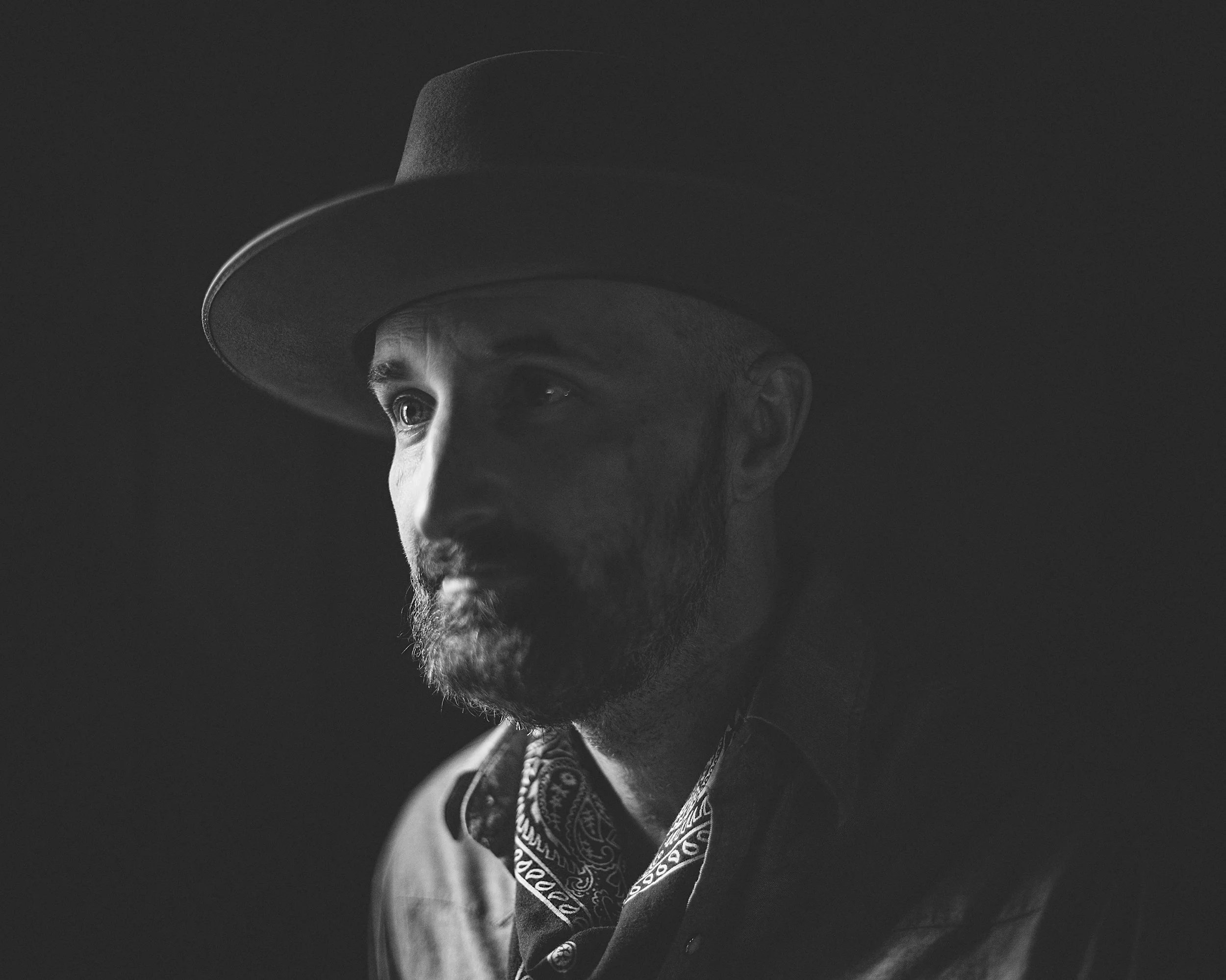 Dramatic black and white studio portrait of blues singer-songwriter Seth Walker wearing a brimmed hat and neckerchief, photographed in 2017 by Parker J. Pfister.