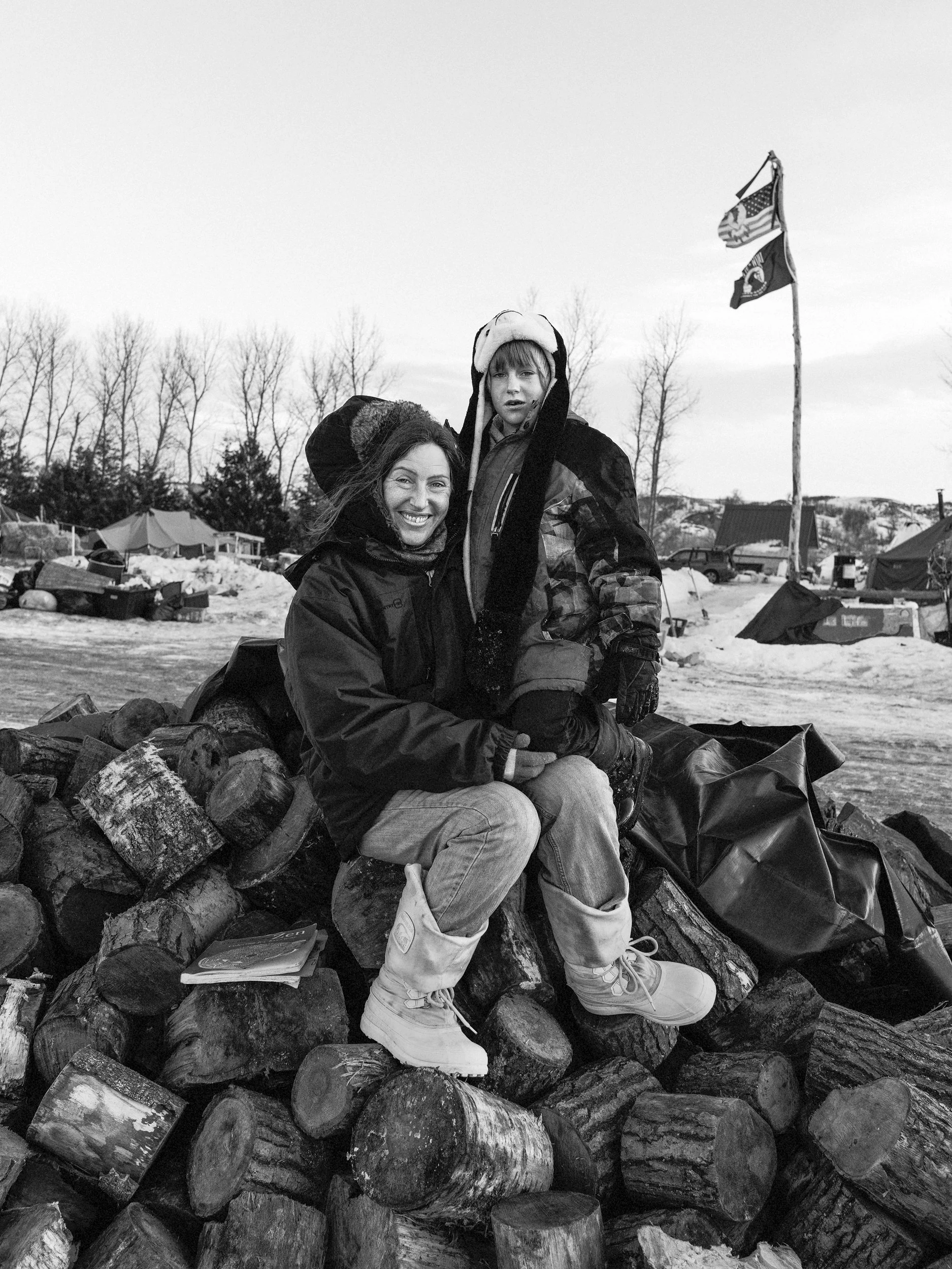 Black and white documentary photograph looking across a snow-covered expanse toward two large flags flying from a flagpole against a pale winter sky at the Standing Rock camp.