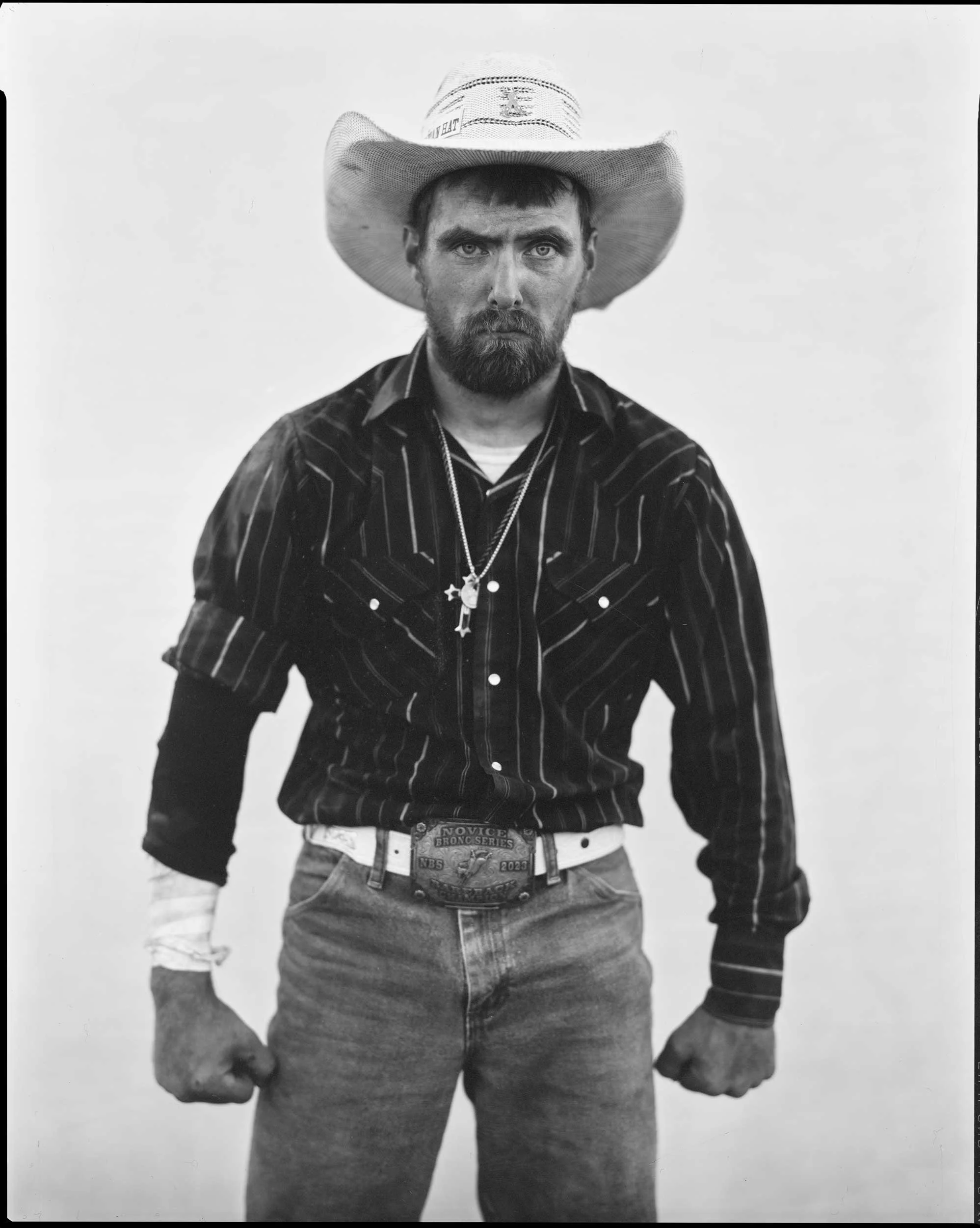 A powerful black and white studio portrait of a bearded man wearing a cowboy hat, western striped shirt, and a large belt buckle, standing with fists clenched against a plain backdrop in Western North Carolina.