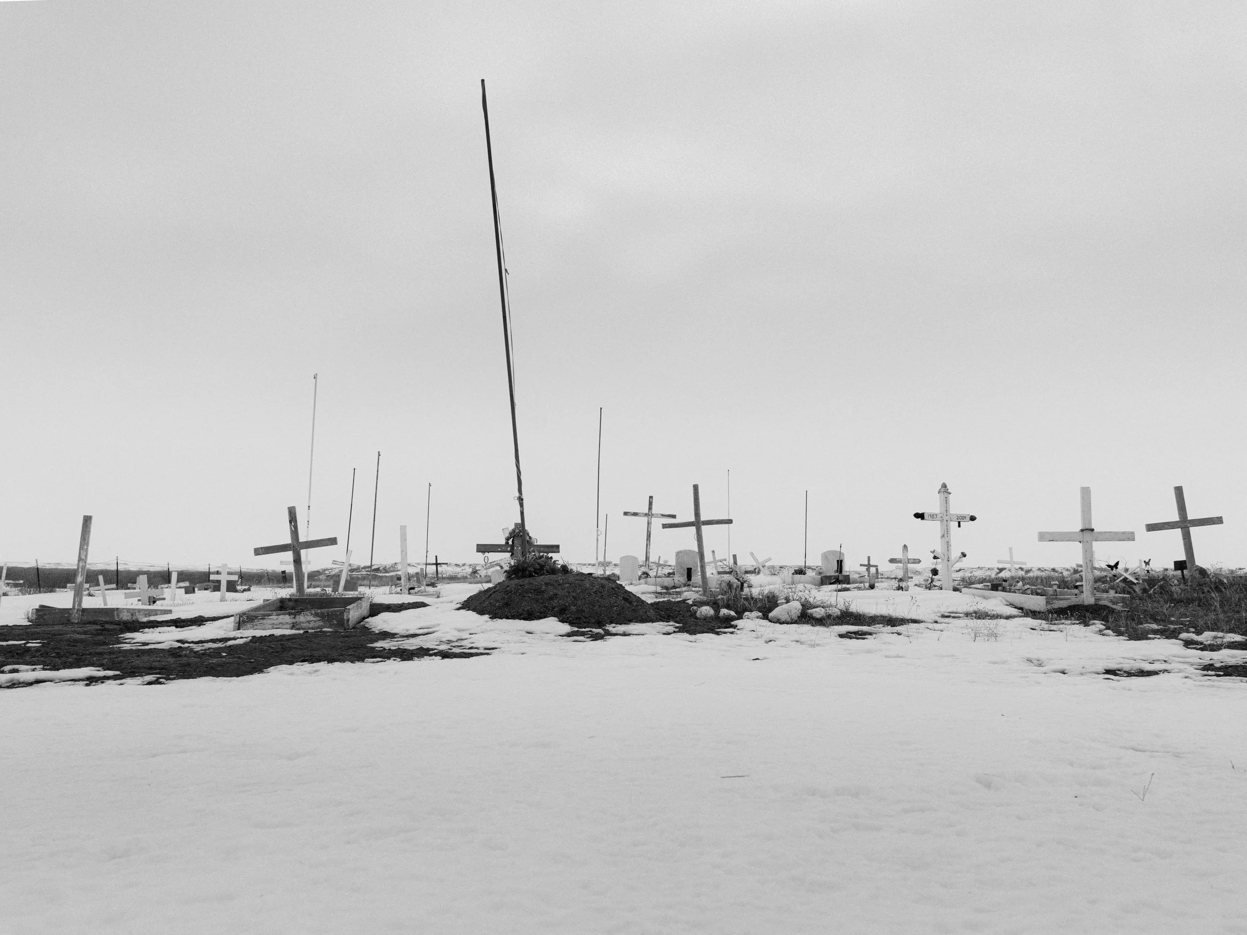 Black and white documentary photograph of a hillside cemetery at the Standing Rock camp covered in snow, featuring numerous simple wooden crosses and a single tall pole standing against an overcast winter sky.