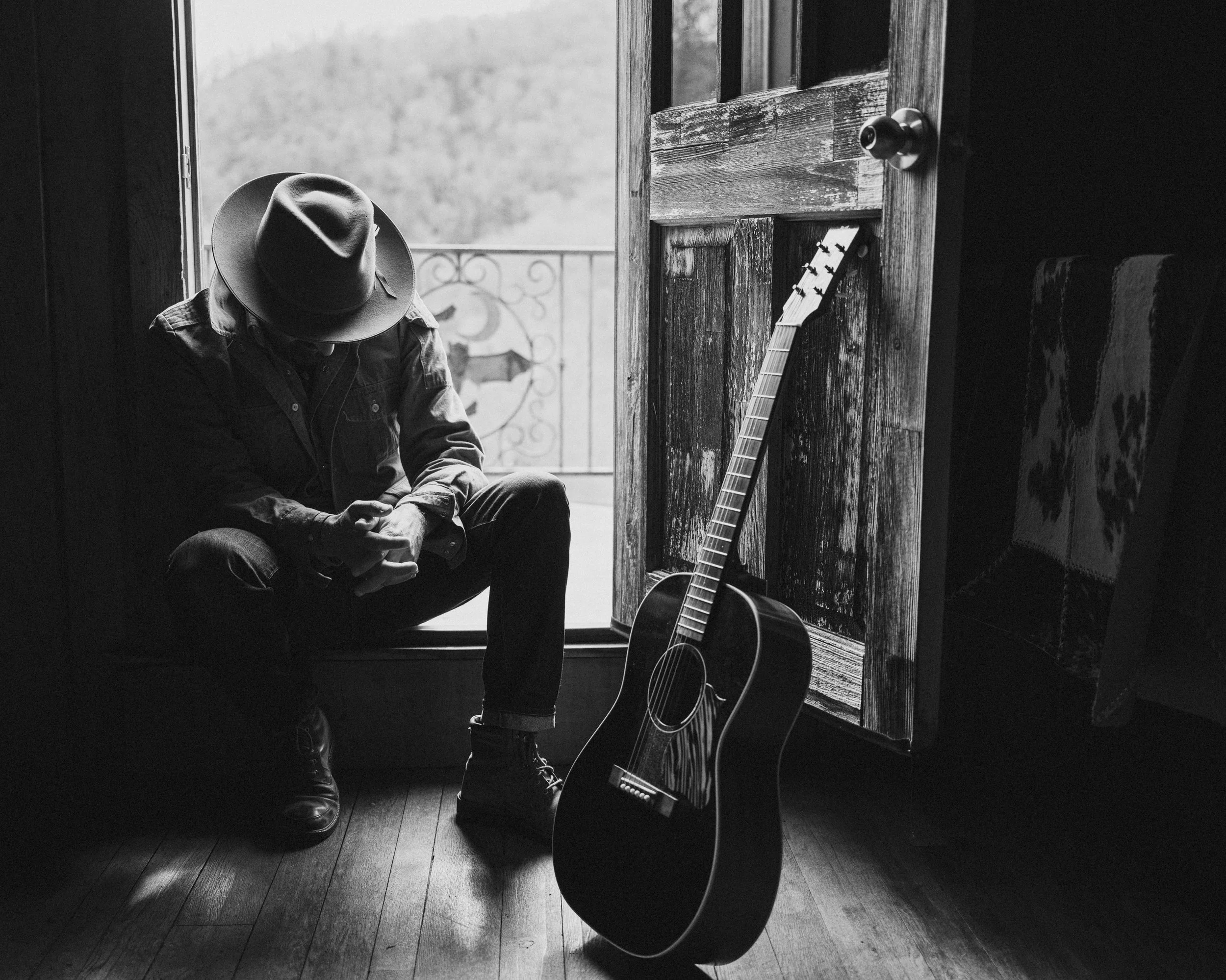 Black and white portrait of blues musician Seth Walker seated in a doorway wearing a brimmed hat next to an acoustic guitar, photographed by Parker J. Pfister.