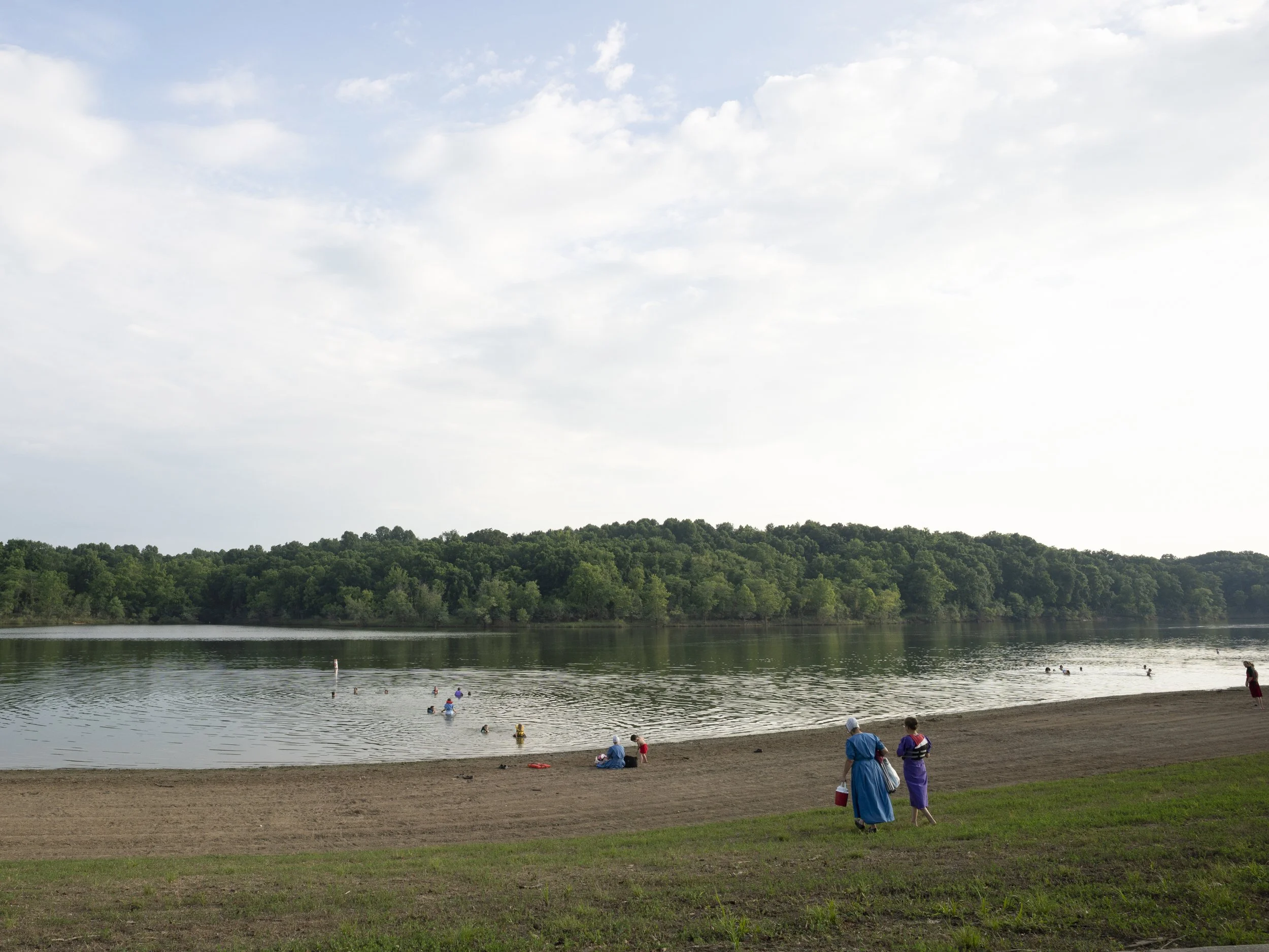 A wide color environmental photograph of Amish or Mennonite women in traditional blue dresses walking along a sandy lake beach, with swimmers in the water and a dense green forest in the background, from One Way Out, Lord by Parker J Pfister.