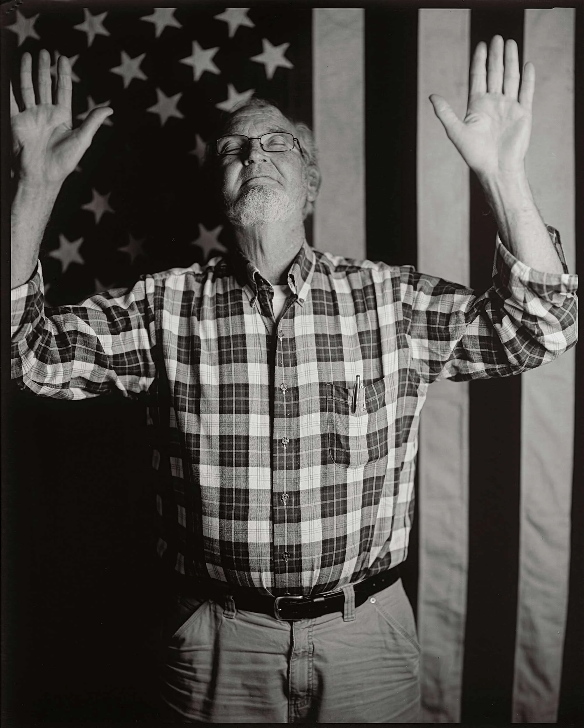 Black and white documentary portrait of an elderly man in a plaid shirt standing with both hands raised, eyes closed, against a large American flag background in Western North Carolina.