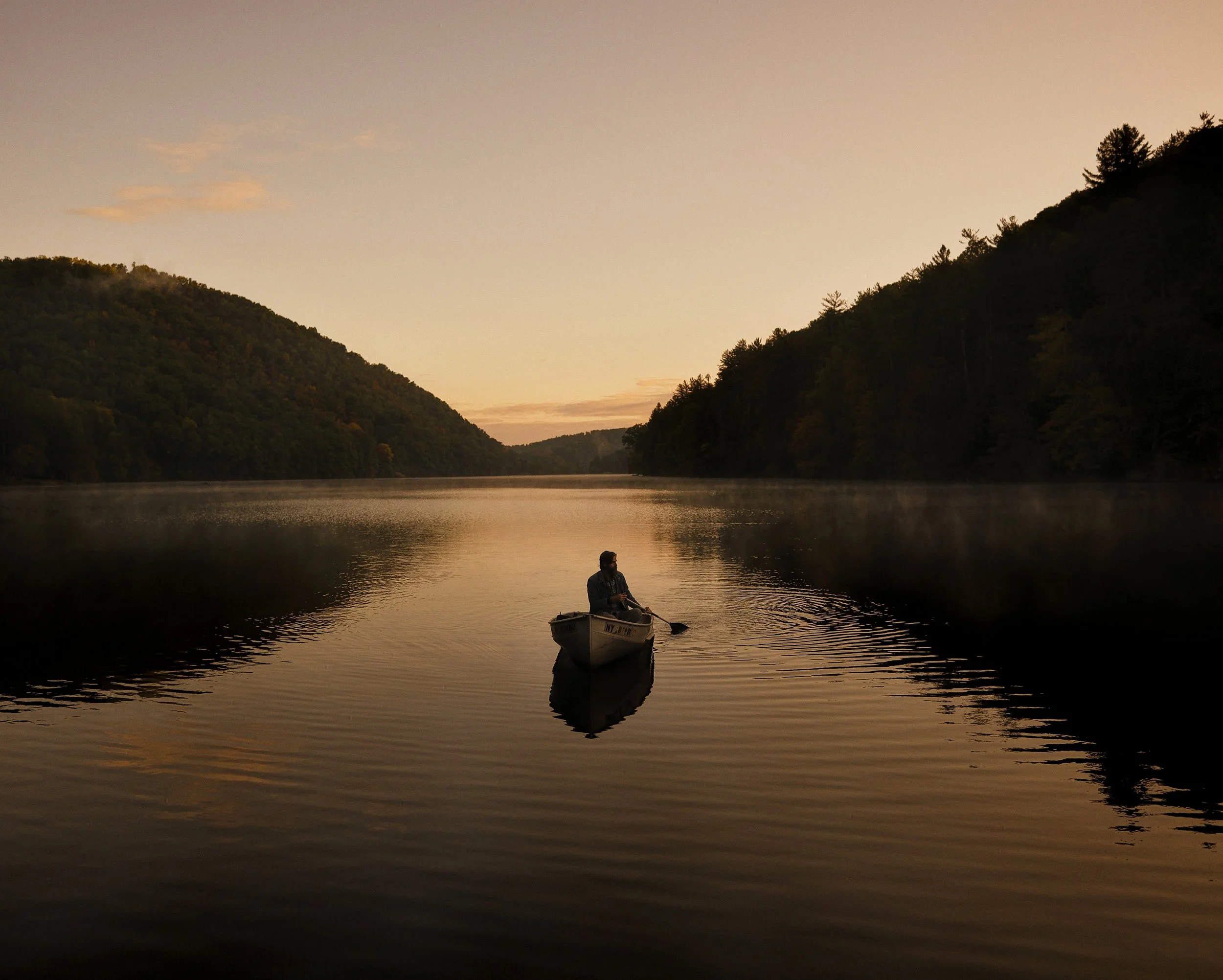 Album cover photograph for New Lost Ages by Tyler Ramsey, featuring a silhouetted figure paddling a canoe at sunset across a calm mountain lake, photographed by Parker J. Pfister.