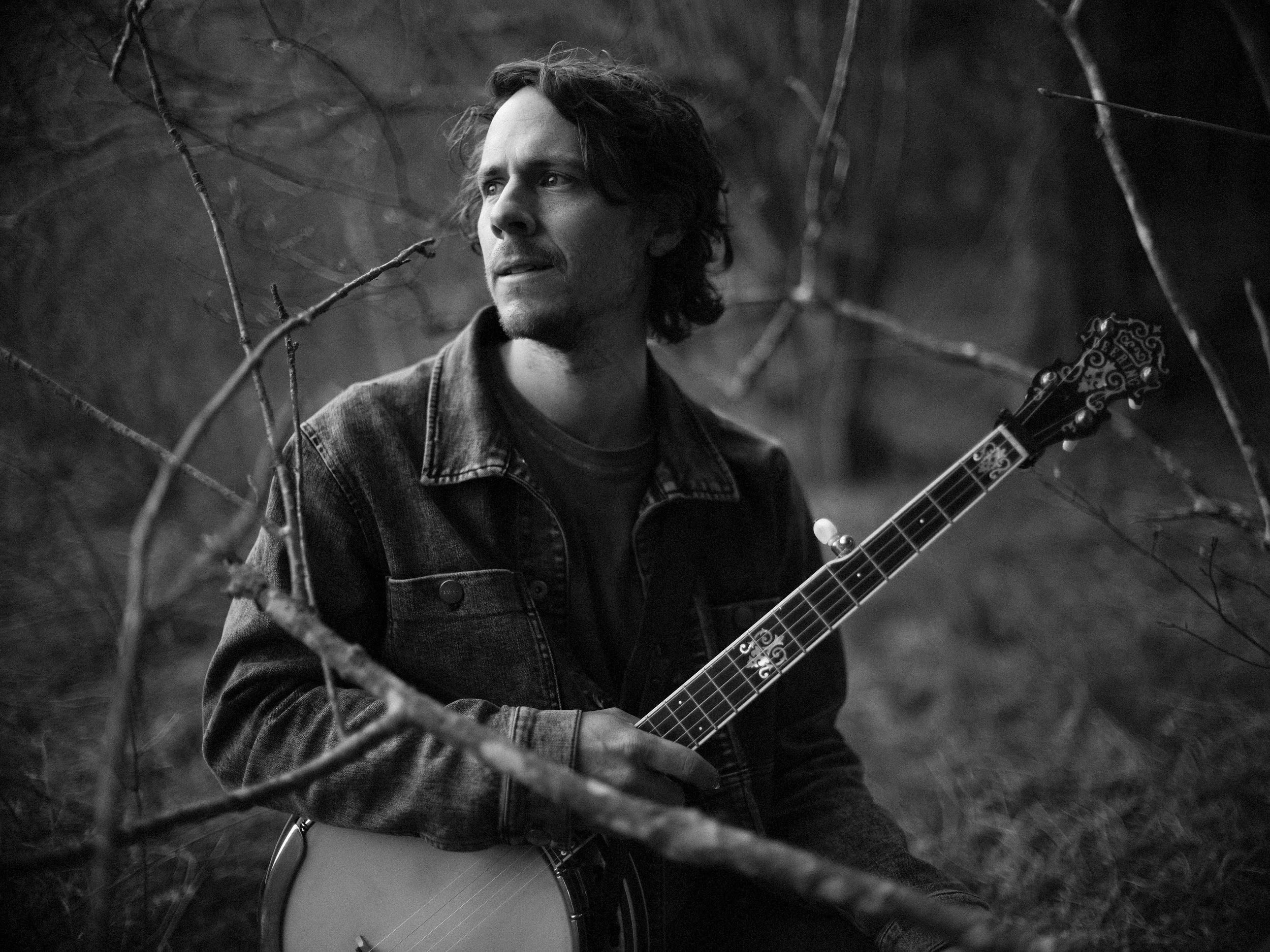 Black and white portrait of banjo player Ross Wall of Western North Carolina band Blue Ridge Betty holding a banjo outdoors among trees, photographed by Parker J. Pfister.