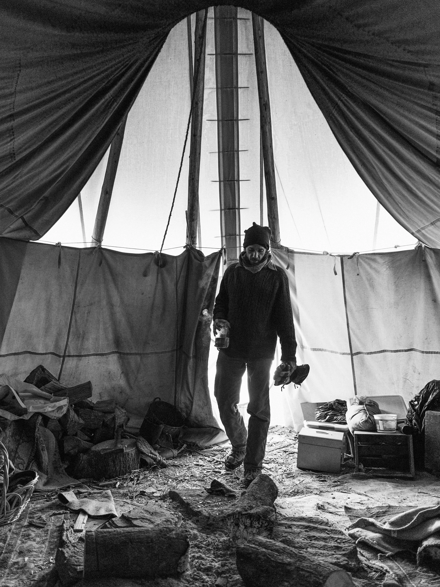 Close-up black and white documentary photograph of the interior floor of a traditional tipi at the Standing Rock camp, showing large cut logs, burlap sacks, and scattered debris on the dirt ground with a person walking in..