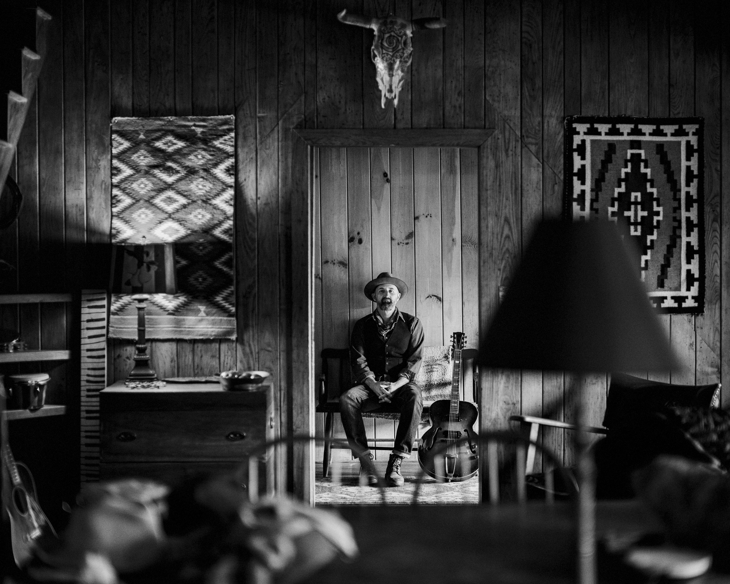 Black and white portrait of blues singer-songwriter Seth Walker seated inside a rustic cabin studio surrounded by guitars, photographed at The Batcave Inn North Carolina by Parker J. Pfister.
