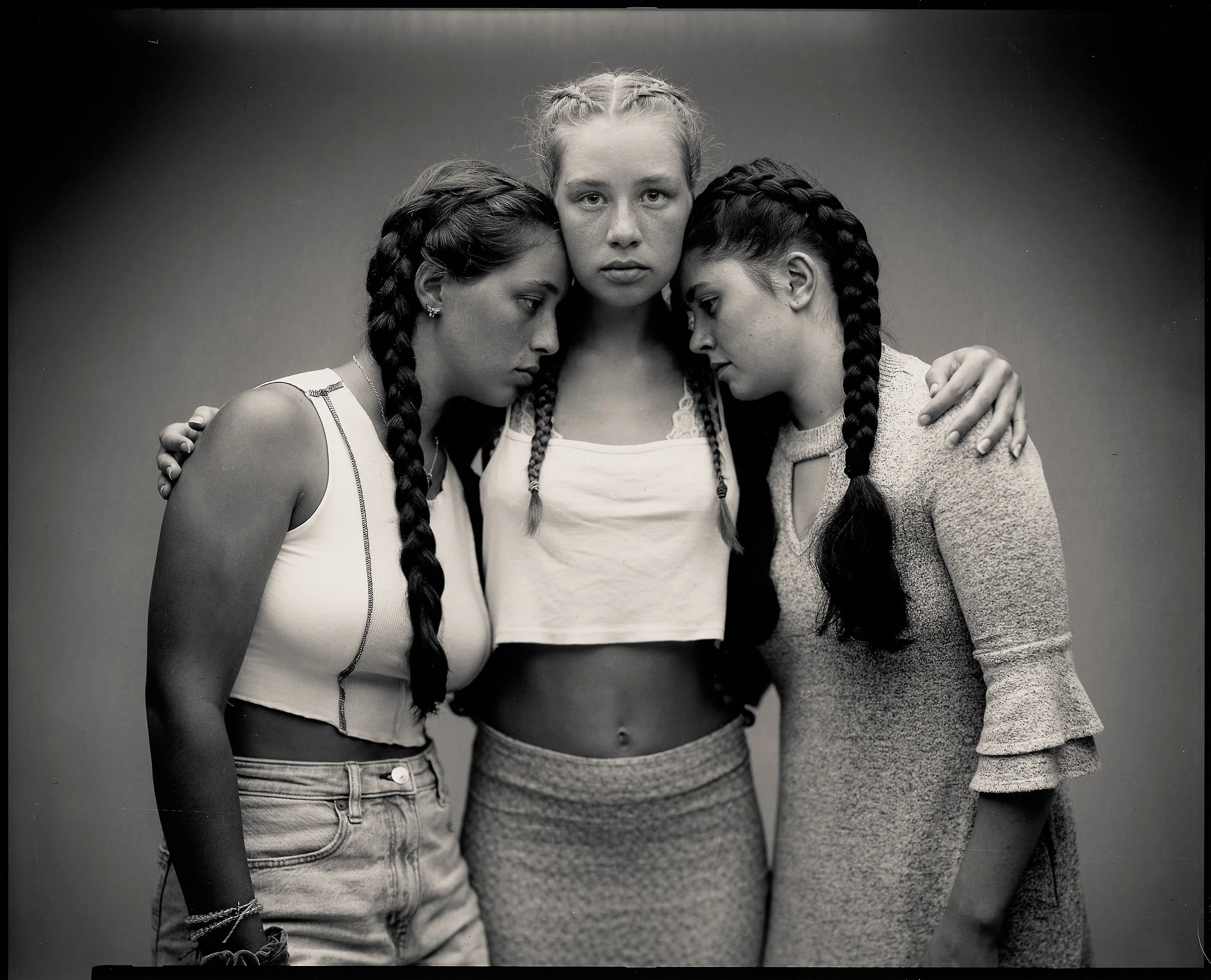 Intimate black-and-white studio portrait of three young people with intricate braided hairstyles, standing close together with gentle, supportive physical contact and thoughtful expressions.