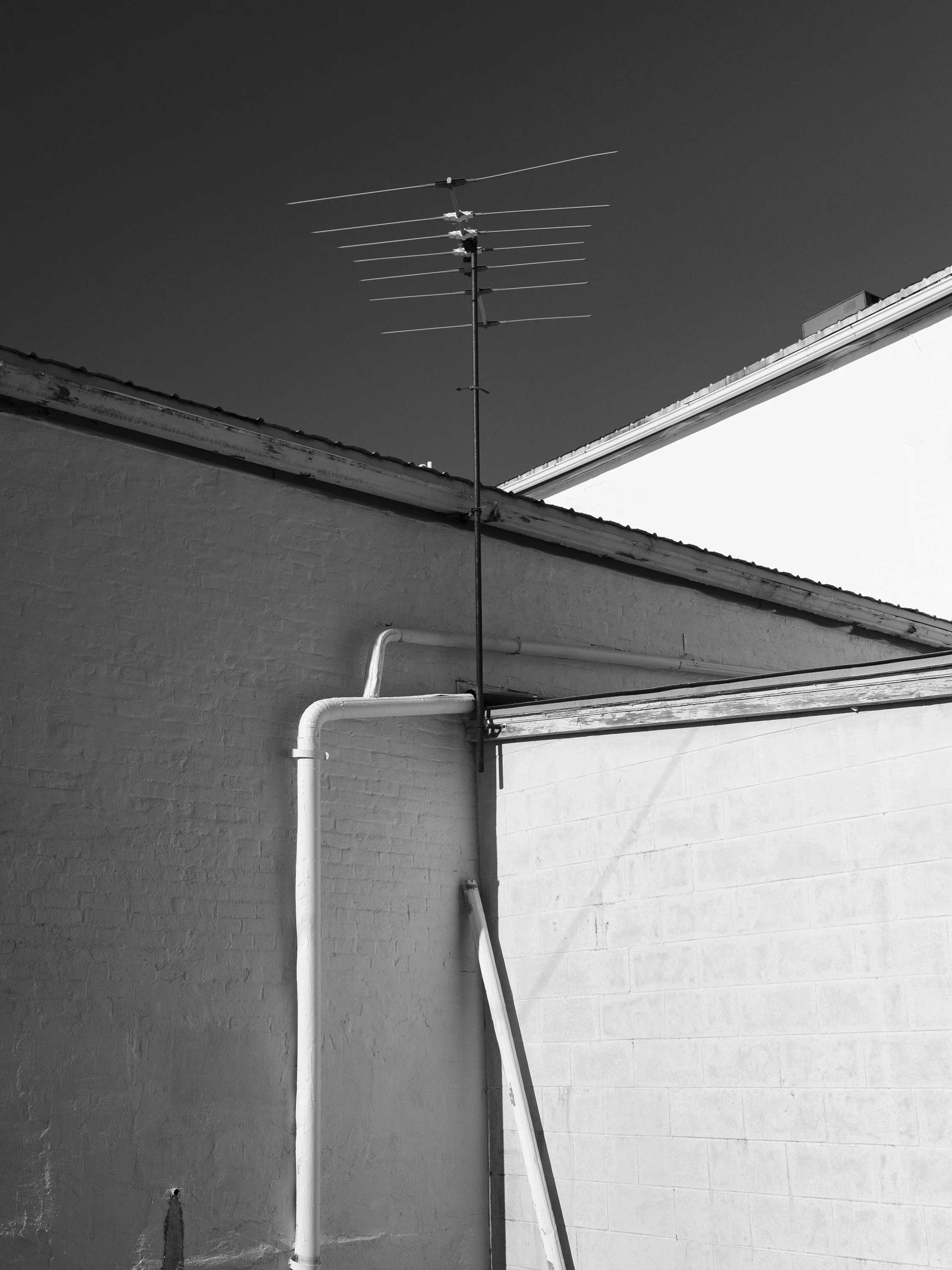 A crisp black and white architectural photograph focusing on white utility pipes and a tall television antenna mounted to the side of a building, from One Way Out, Lord by Parker J Pfister.