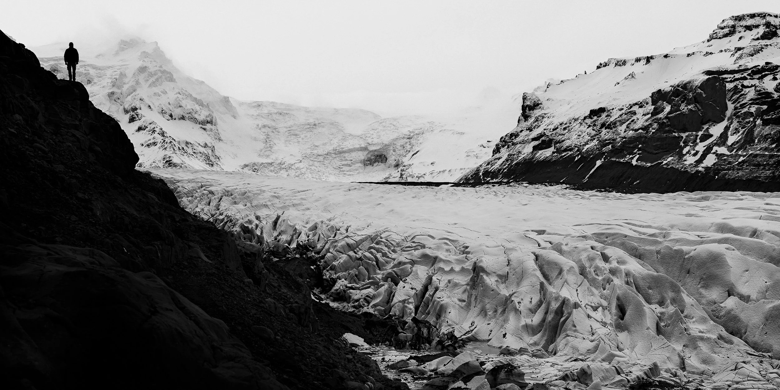 A sweeping black and white fine art photograph of a massive, heavily textured glacier valley in Iceland. A lone silhouette of a person stands high on a dark, steep rocky ridge on the left, looking out over the vast, rugged expanses of glacial ice.