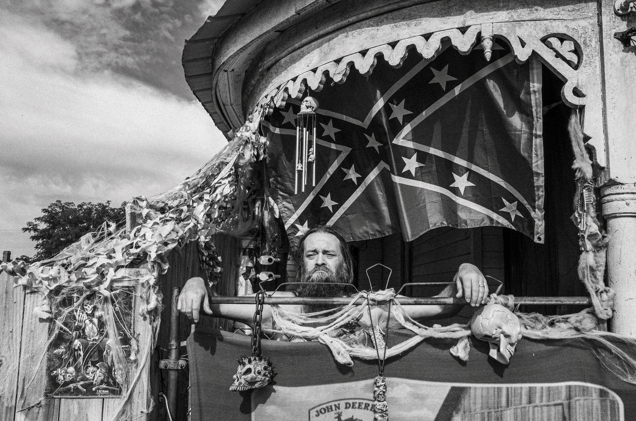 A detailed black and white environmental portrait of a bearded man leaning over a porch heavily decorated with skulls, netting, a John Deere banner, and Confederate flags, from the One Way Out, Lord documentary series by Parker J Pfister.