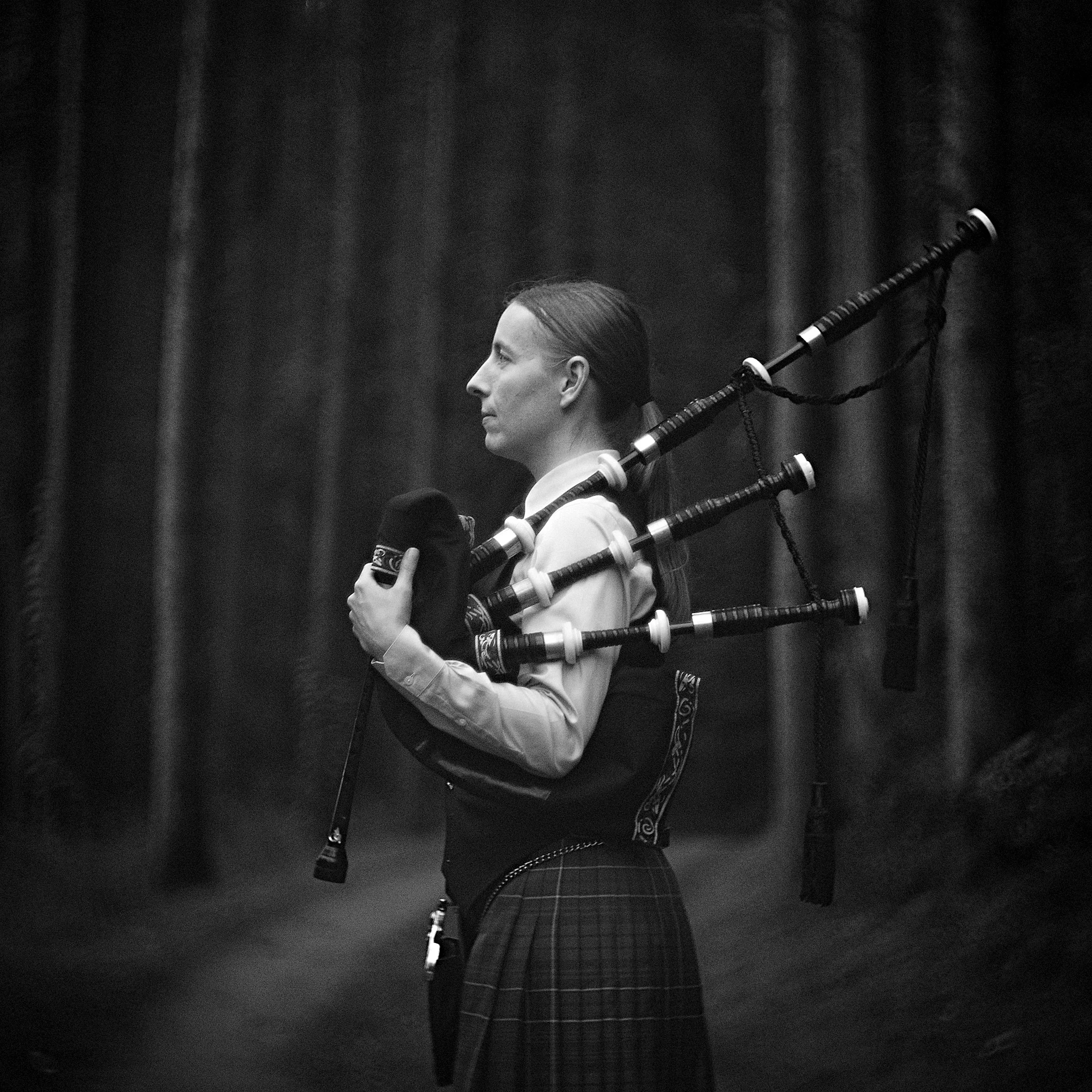 Side-profile fine art portrait of a female bagpipe player in traditional kilt dress standing among tall pines in Candler, North Carolina, photographed by Parker J. Pfister.