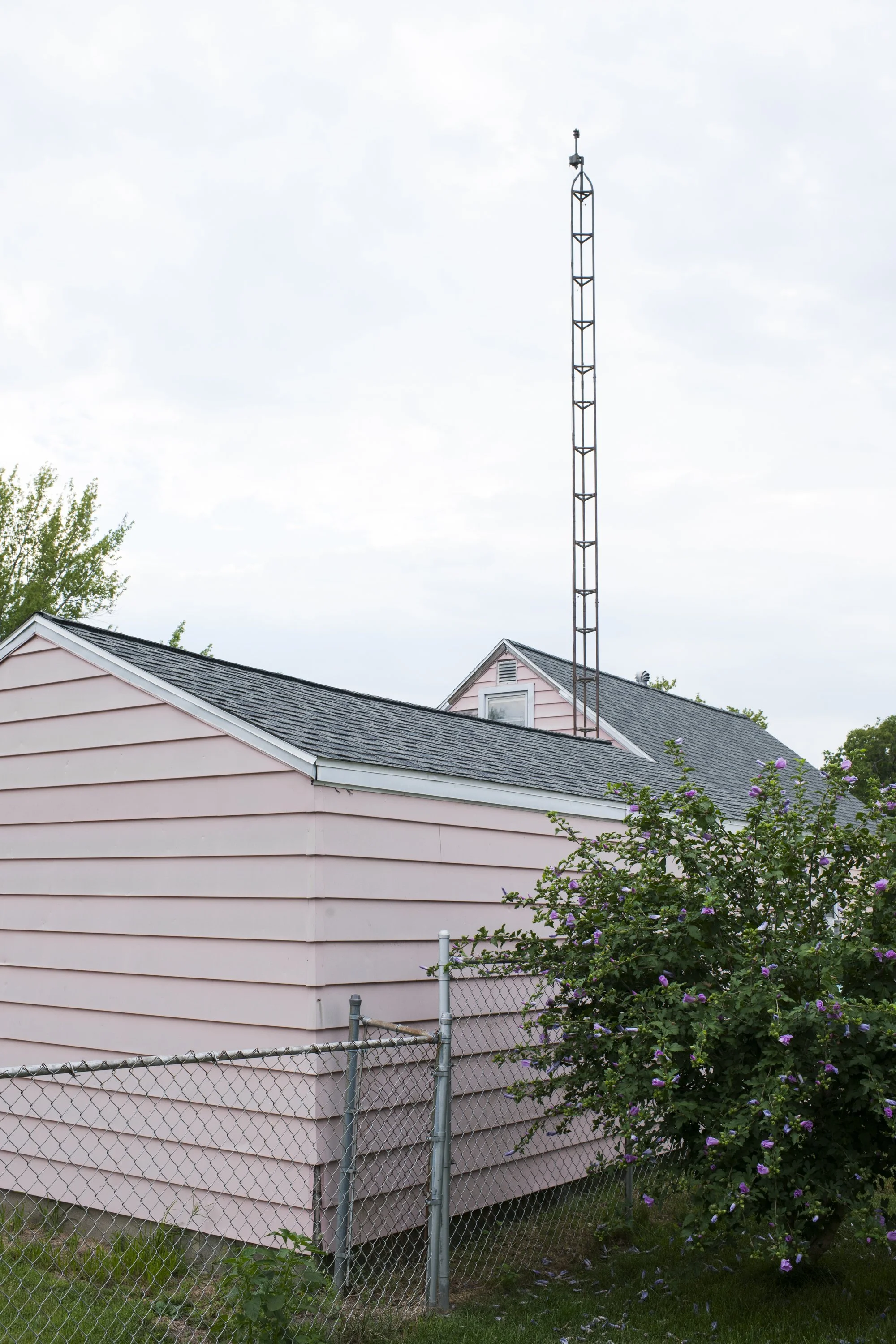 A vertical color photograph of a light pink sided residential building behind a chain-link fence, with a tall television antenna reaching into an overcast sky, from One Way Out, Lord by Parker J Pfister.