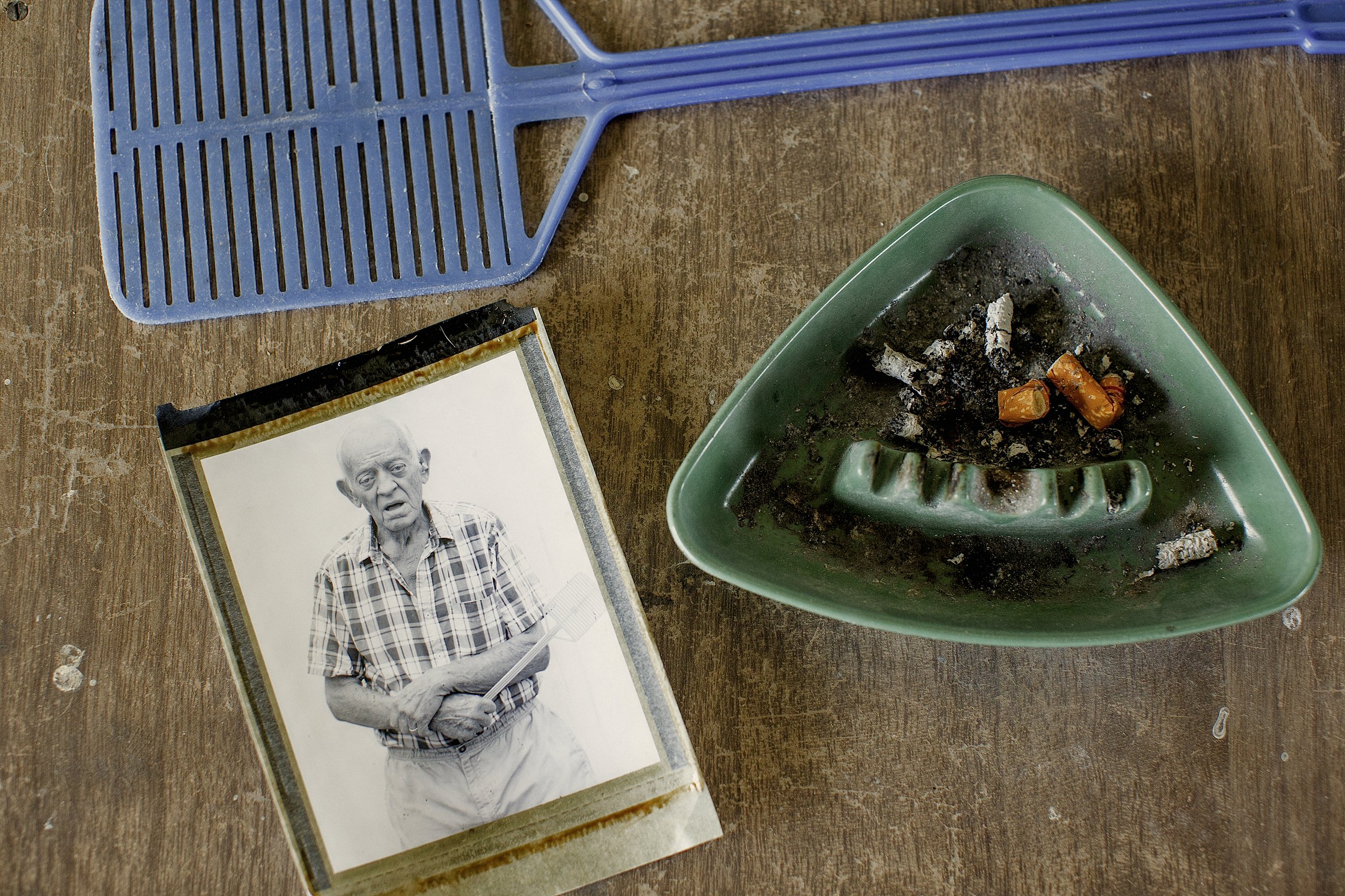 A color film photograph capturing a Type 55 Polaroid print of an older man sitting on a rustic wooden table next to a green triangular ashtray and a blue plastic fly swatter, from One Way Out, Lord by Parker J Pfister.