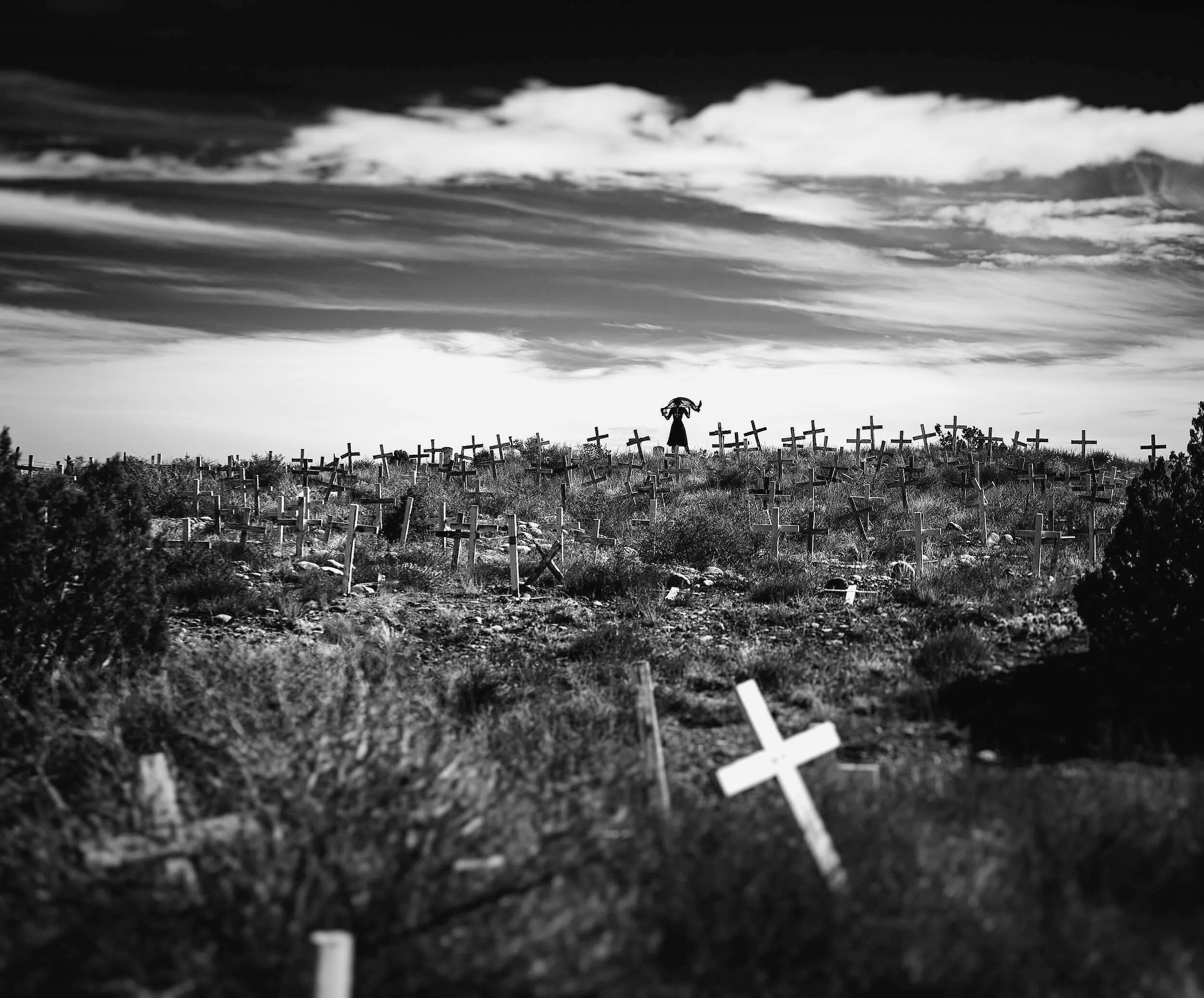 A black and white fine art photograph of a rolling hillside cemetery in New Mexico covered with dozens of simple wooden crosses, with a lone silhouette of a person standing on the horizon under a dramatic, cloudy sky.