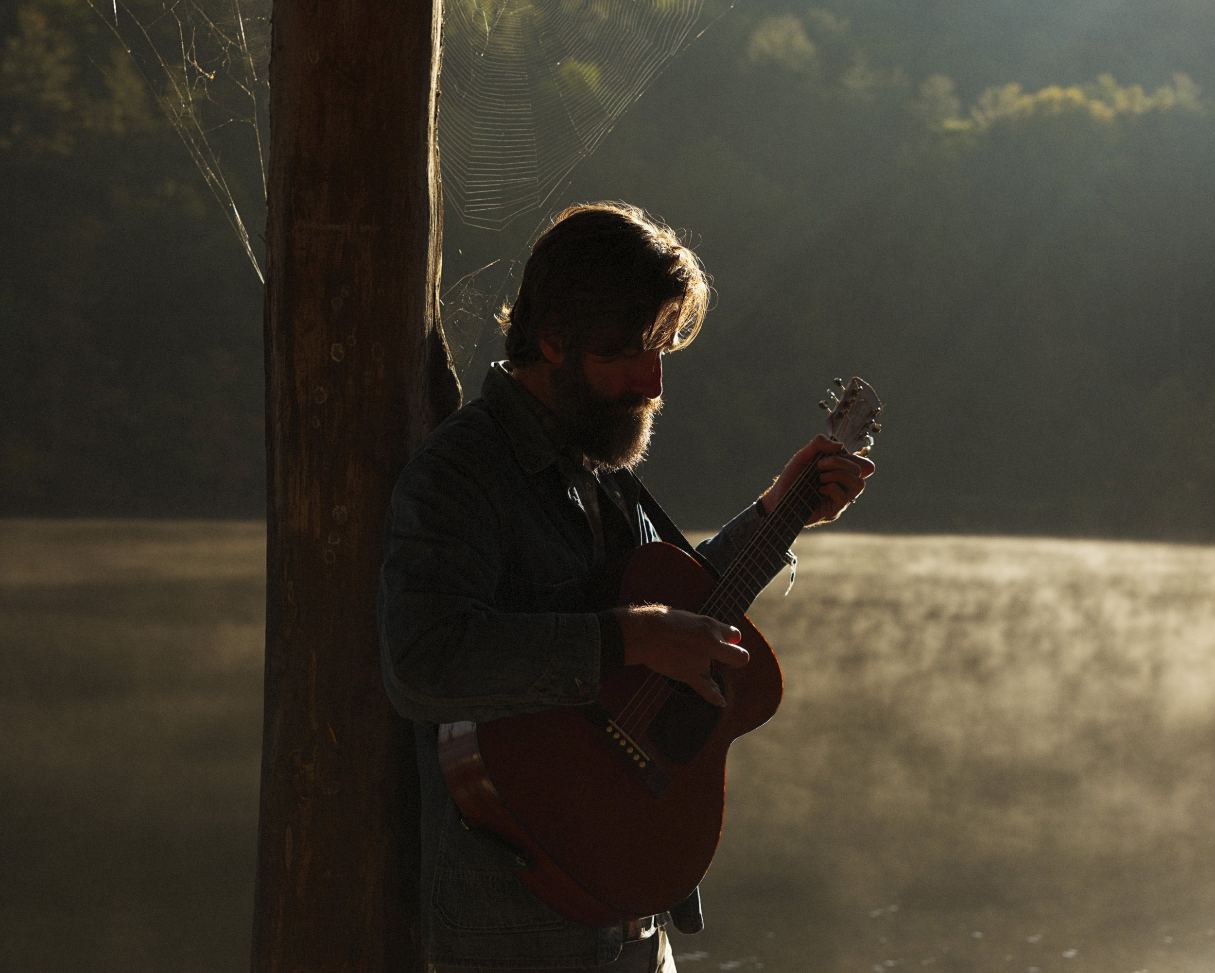 Warm atmospheric portrait of musician Tyler Ramsey playing acoustic guitar beside a tree along a misty lake shoreline, captured for his album New Lost Ages, photographed by Parker J. Pfister.