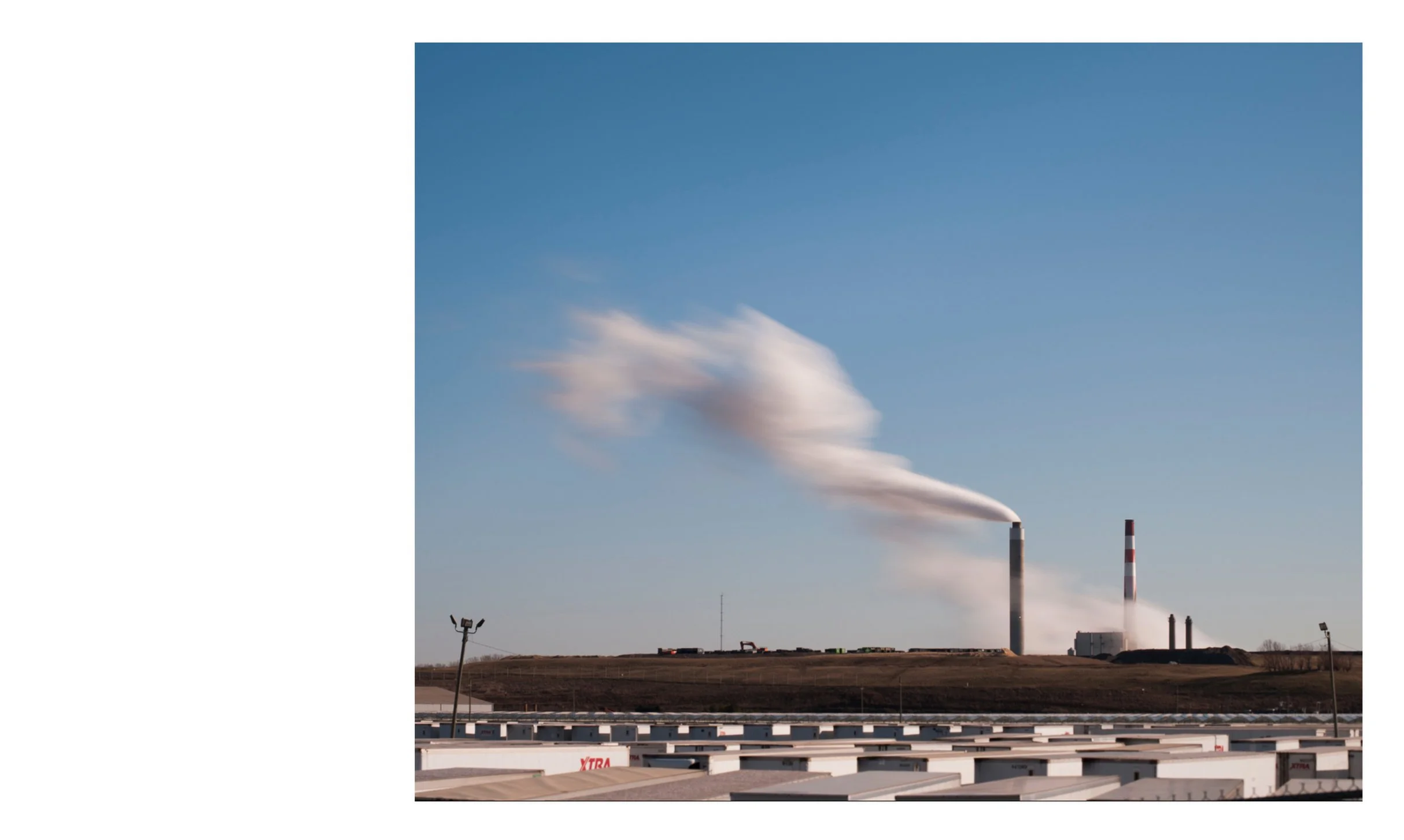 A color documentary photograph featuring a long exposure of white smoke billowing from industrial smokestacks into a blue sky above a line of shipping trailers, from the Connecting Hwy 80 project by Parker J Pfister.