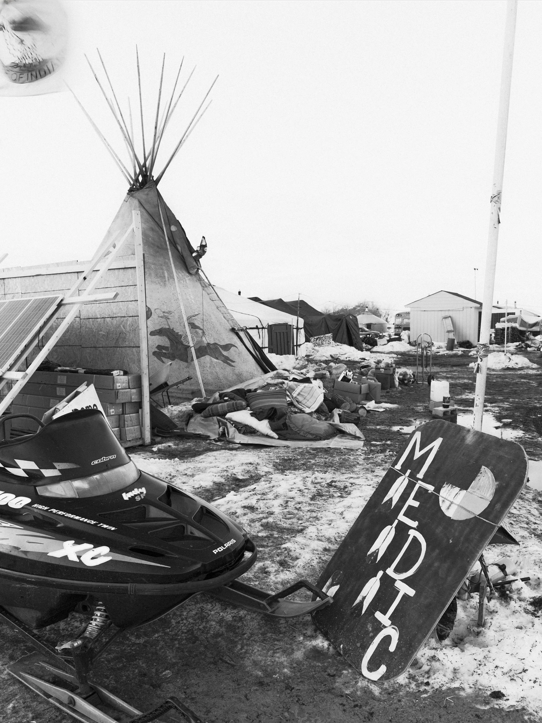 Close-up black and white documentary photograph showing the front skis of a Polaris Indy XC snowmobile parked on patchy snow next to a handmade wooden sign painted with indigenous symbols at the Standing Rock camp.