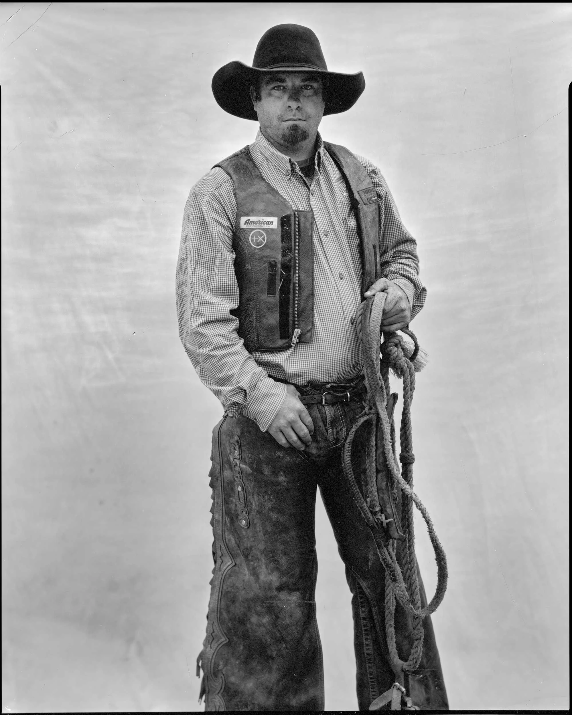 An 8x10 large format black and white paper negative portrait of a bearded man wearing a cowboy hat, leather vest, and chaps, standing against a light backdrop while holding a coiled lasso rope at the Marshall rodeo in Western North Carolina.
