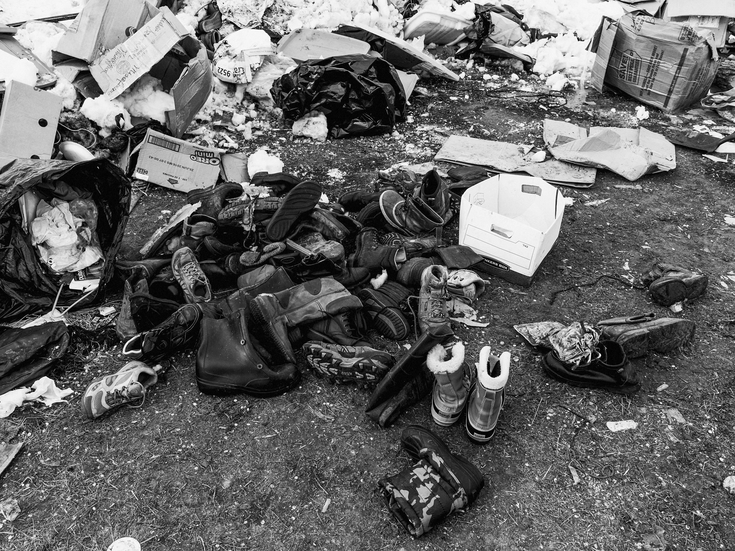 Black and white documentary photograph capturing a large, cluttered pile of winter boots, shoes, and clothing discarded on a patch of muddy, snow-dusted ground amidst flattened cardboard boxes and tarps at the Standing Rock camp.