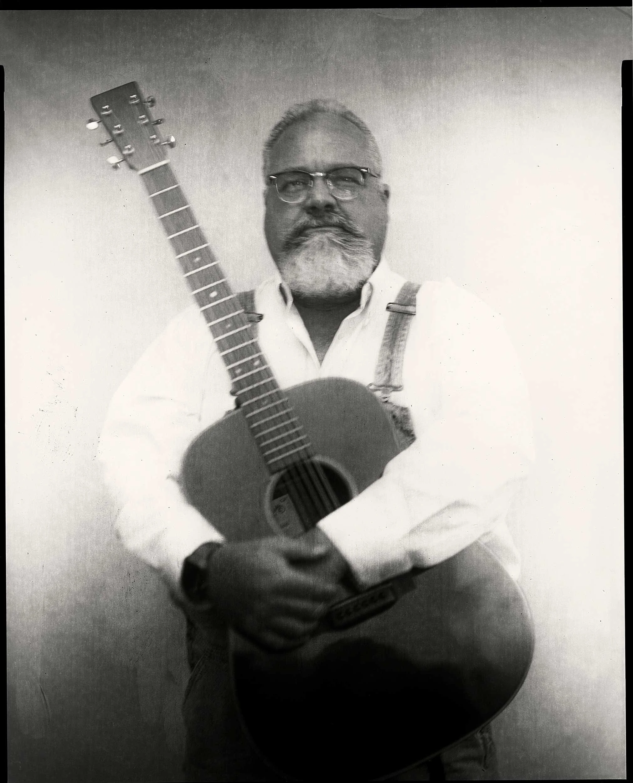 An 8x10 large format black and white paper negative portrait of a smiling, bearded man with glasses wearing overalls and a white shirt, holding an acoustic guitar at the Bluff Mountain Music Festival in Hot Springs, North Carolina.