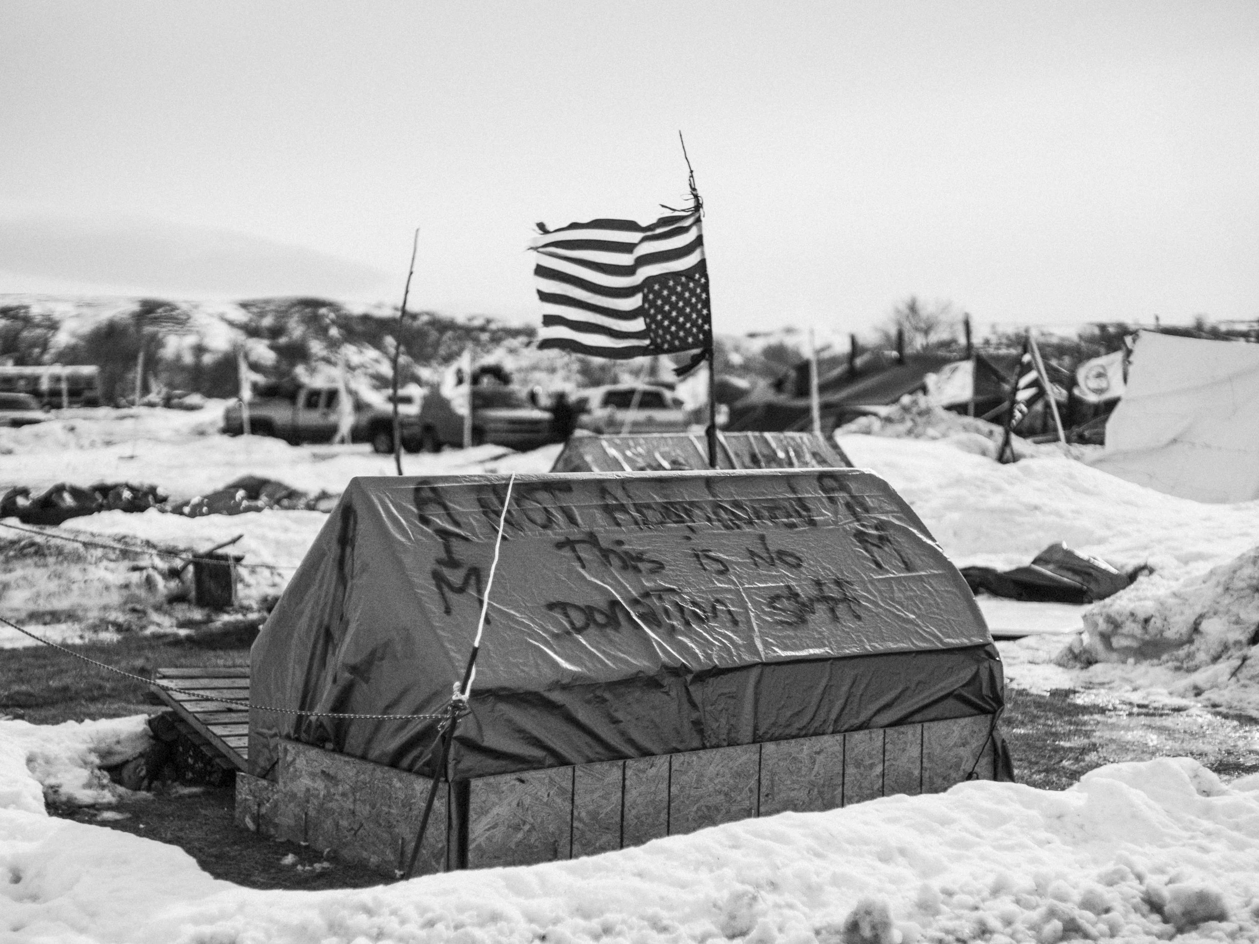 Black and white photograph looking across a vast, flat snow-covered landscape at the Standing Rock camp under an overcast winter sky.