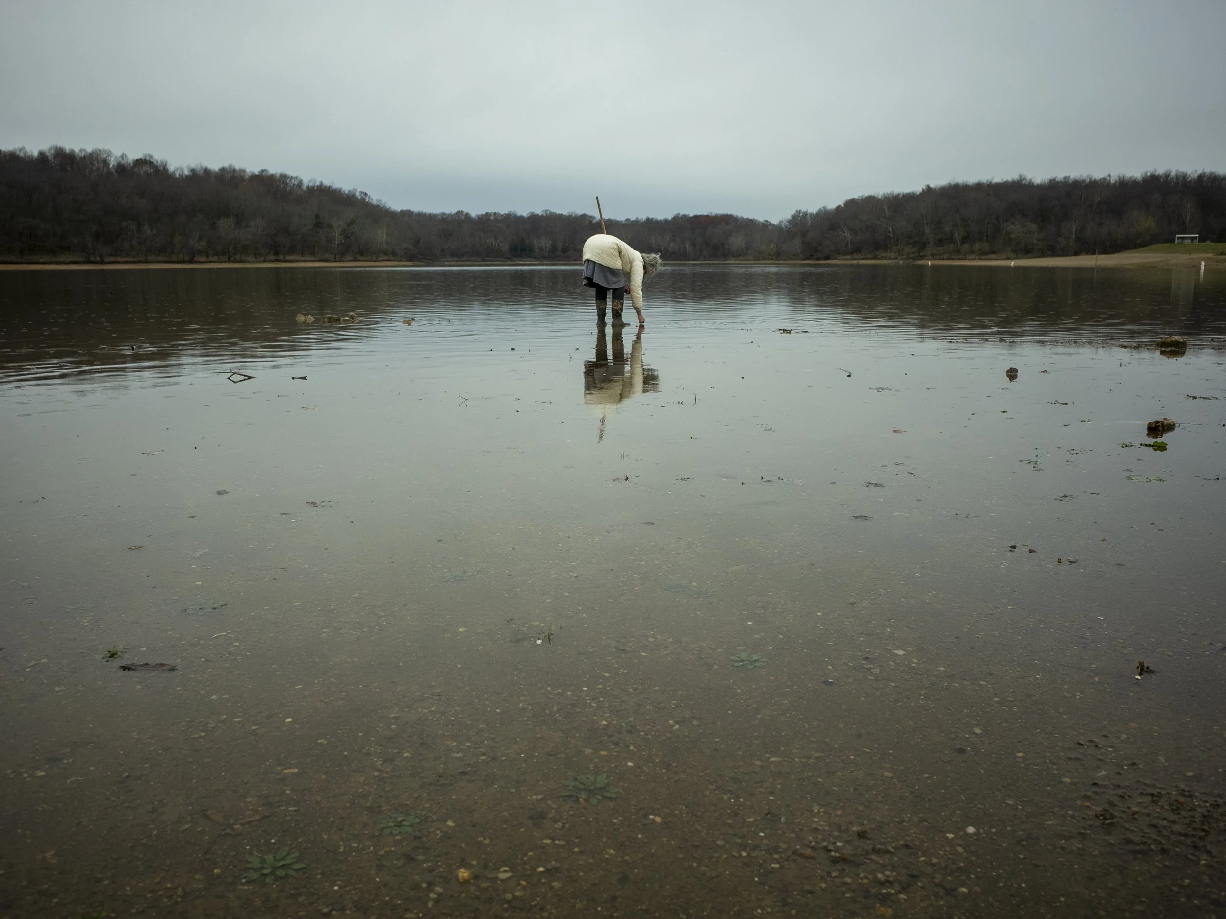 A color documentary photograph of a woman wading into a calm, reflective lake under an overcast sky, bending down to search the shallow water for fossils, from One Way Out, Lord by Parker J Pfister.